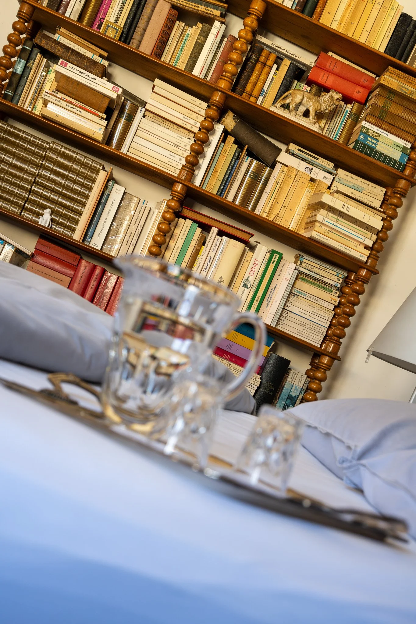 Une étagère en bois contenant de nombreux livres de différentes tailles et couleurs, avec des décorations telles qu'une statue de lion et une petite figurine, vue de côté dans une pièce à la lumière chaude.