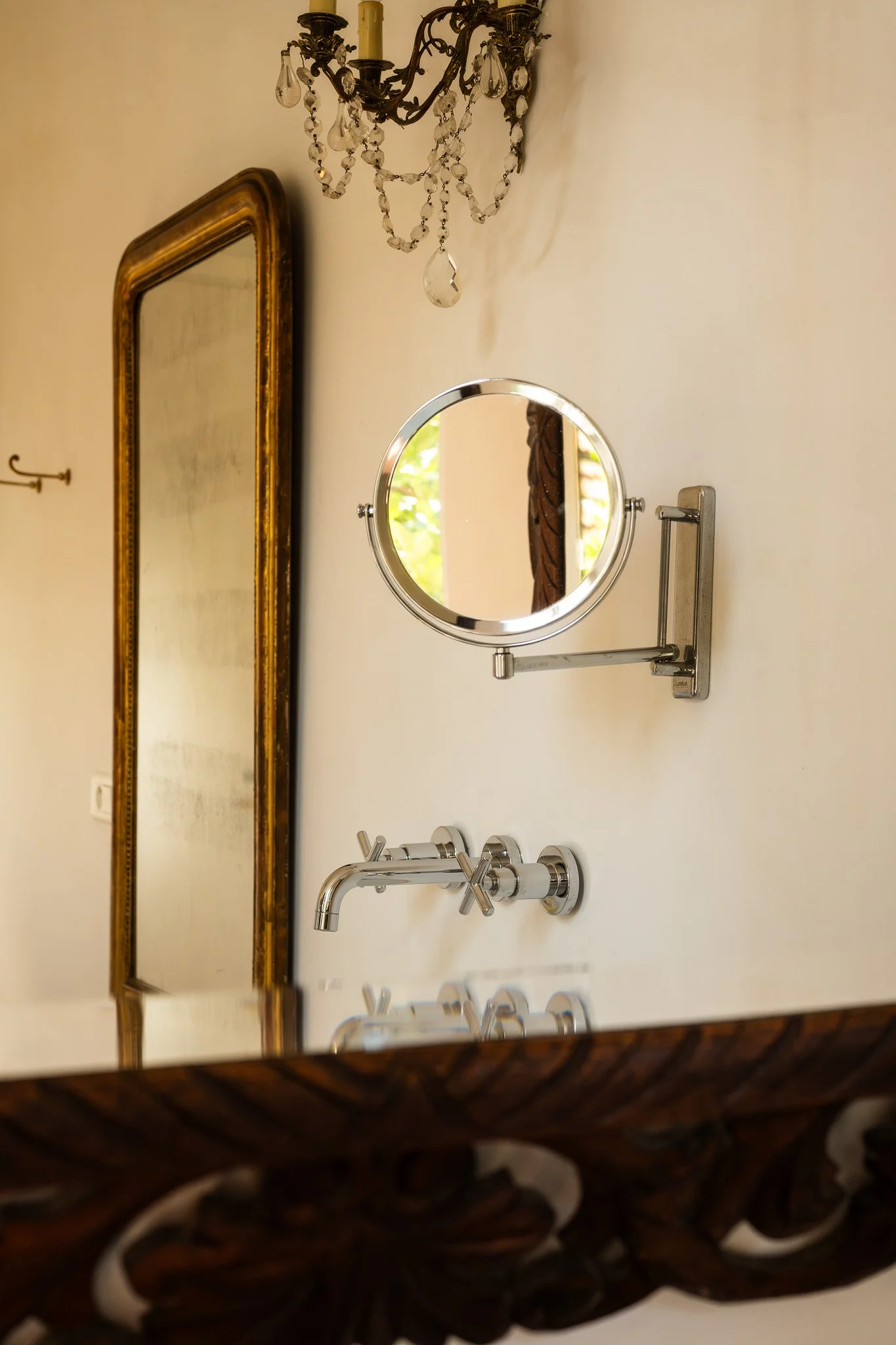 Un miroir rond avec une base métallique, une lampe en cristal suspendue au plafond, une grande fenêtre avec rideau et une table avec des robinets en dessous.
