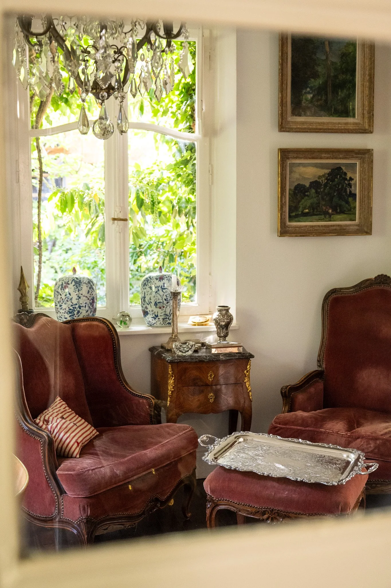 Intérieur d'un salon élégant avec fauteuils en velours rouge, une table en marbre marron, des décorations en argent, un chandelier en cristal suspendu, et des tableaux représentant des paysages, avec une grande fenêtre laissant entrer la lumière naturelle et laissant voir la verdure extérieure.