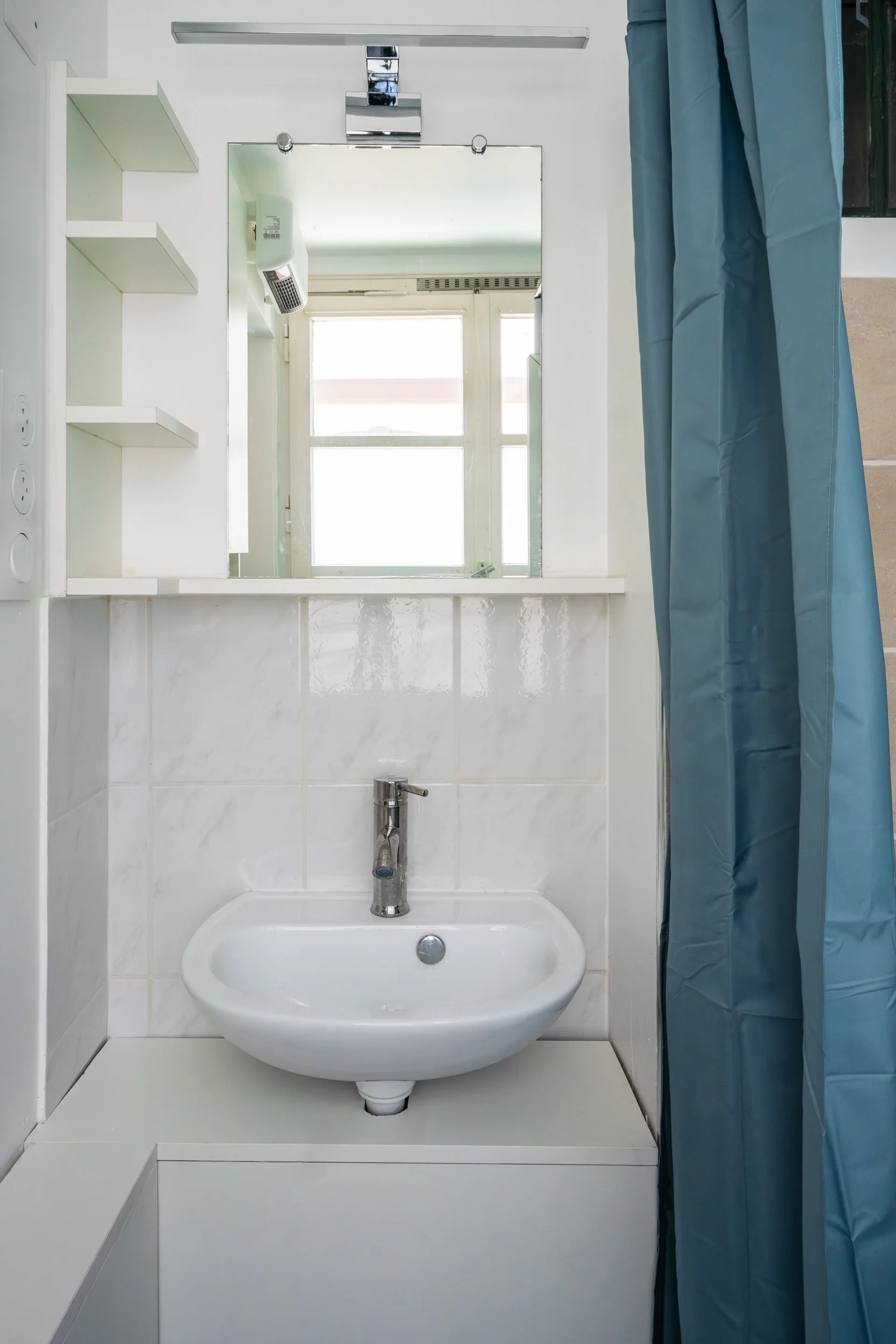Vue d'une salle de bain blanche avec un lavabo, un miroir, un radiateur, un distributeur de savon et une fenêtre, avec un rideau bleu à droite.