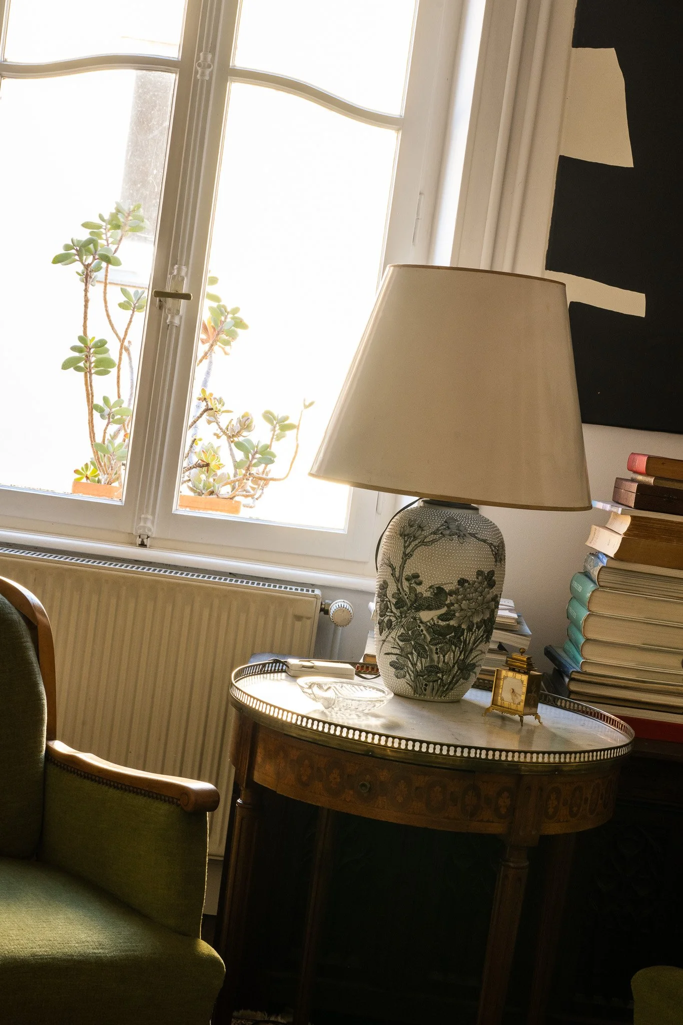 Intérieur d'un salon avec une table ronde en bois, une grande lampe en porcelaine avec un motif floral, une pile de livres, une fenêtre laissant entrer la lumière, un canapé vert, et un tableau noir suspendu au mur.