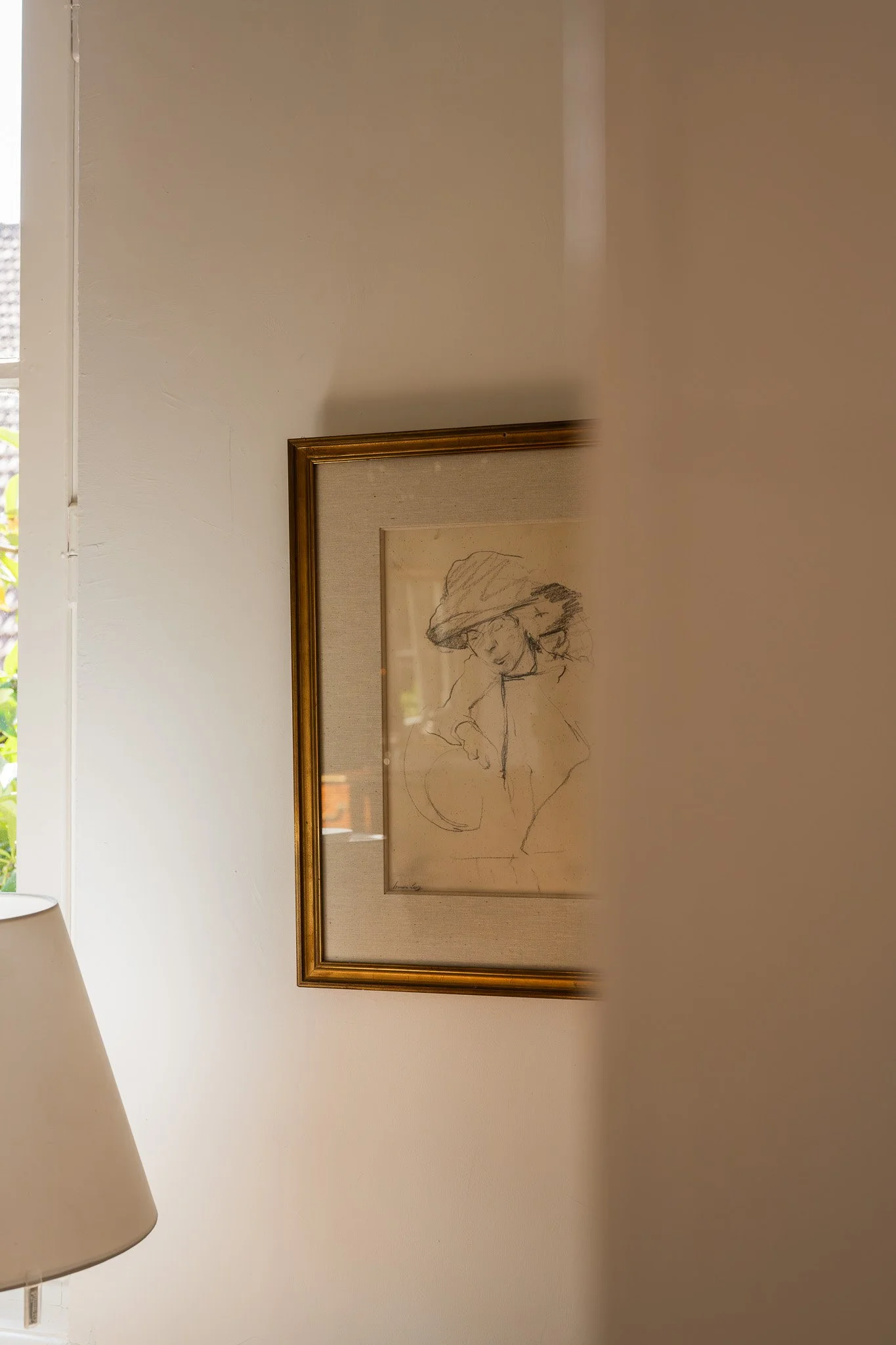 Un mur blanc avec une peinture encadrée représentant un dessin d'une femme portant un chapeau. Une lampe beige est visible en bas à gauche.