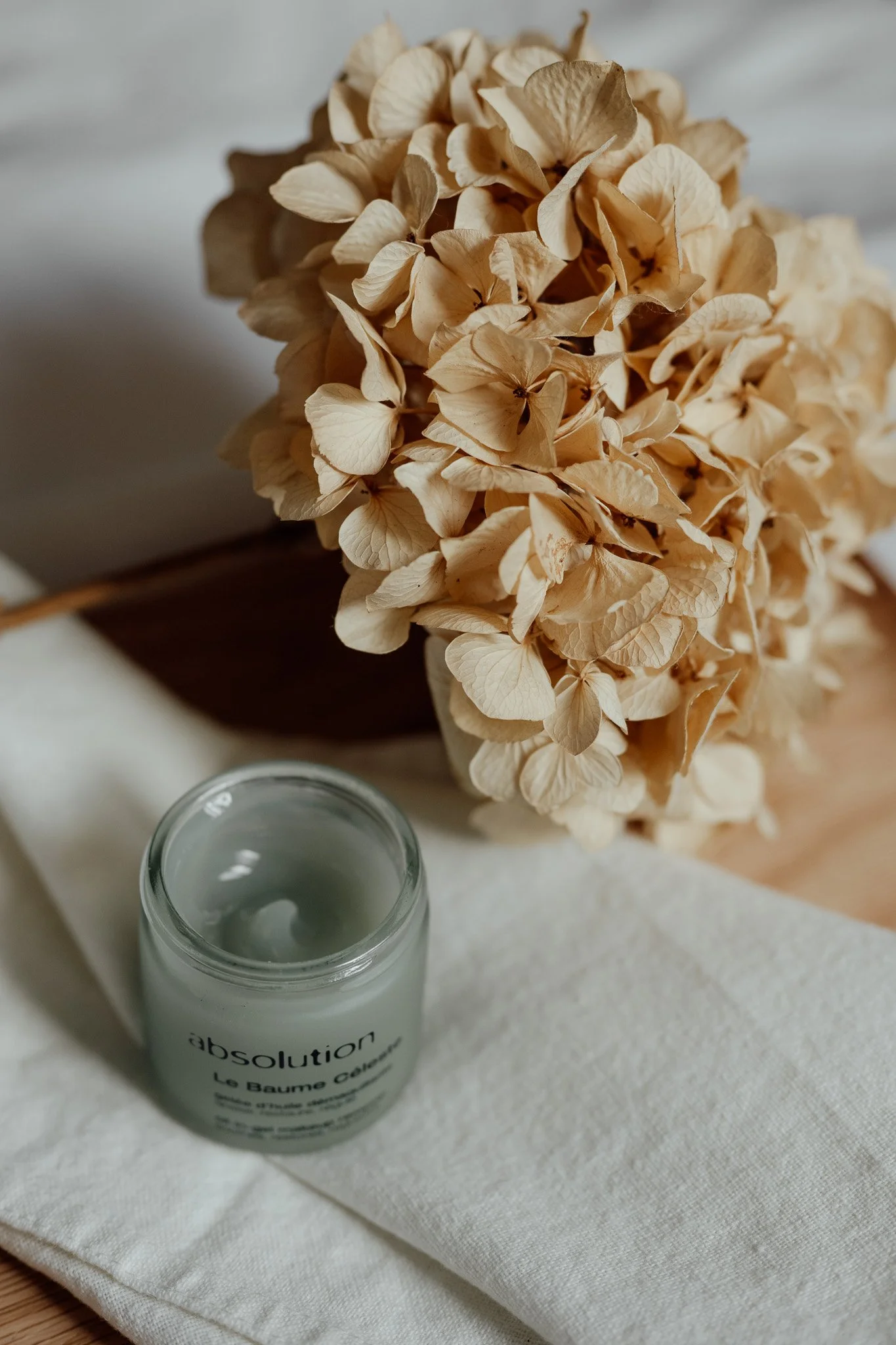 Un bouquet de fleurs séchées beige et un petit pot de crème dans un contenant en verre blanc sur une nappe en tissu blanc.