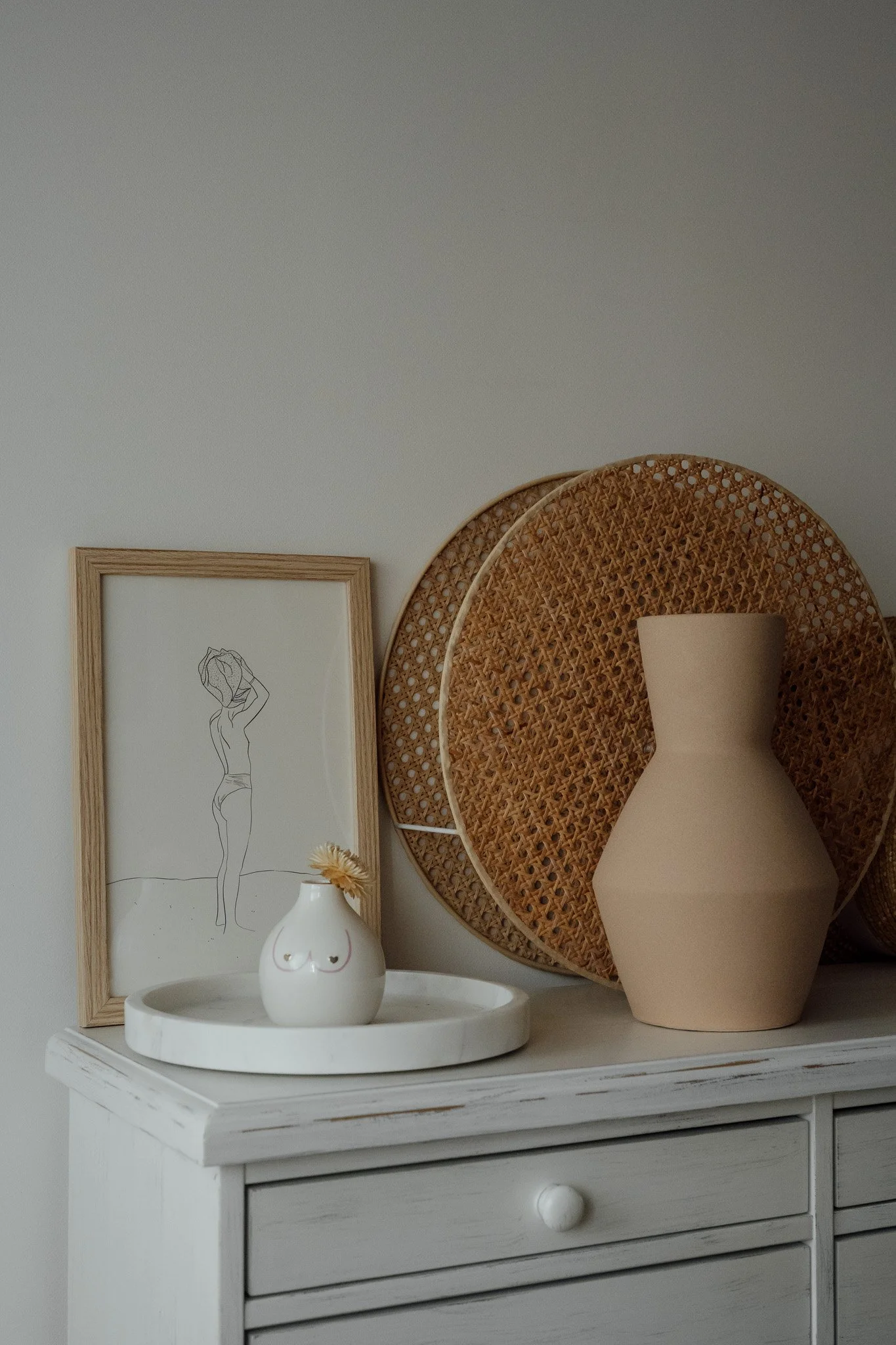 Une composition de décoration intérieure avec un tableau avec une illustration minimaliste d'une femme, deux grands paniers en rotin, une lanterne beige et une petite boule blanche avec un visage souriant, tous placés sur une commode blanche.