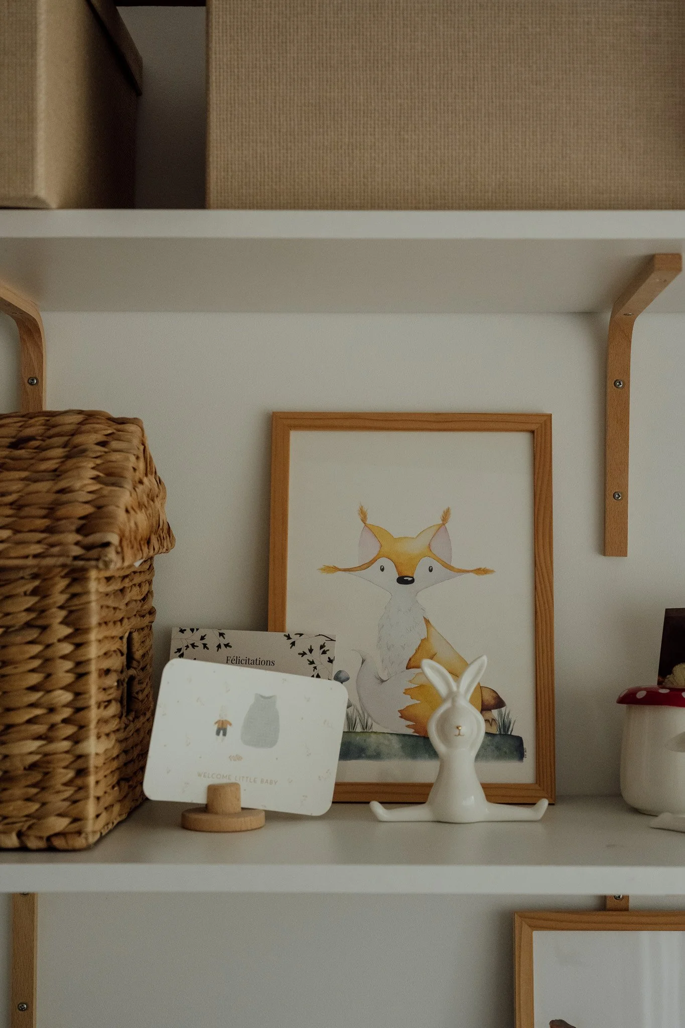 Une étagère avec des boîtes en carton, une illustration d'un renard en dessous d'une photo avec un cadre en bois, une figurine de lapin blanche et une tasse.