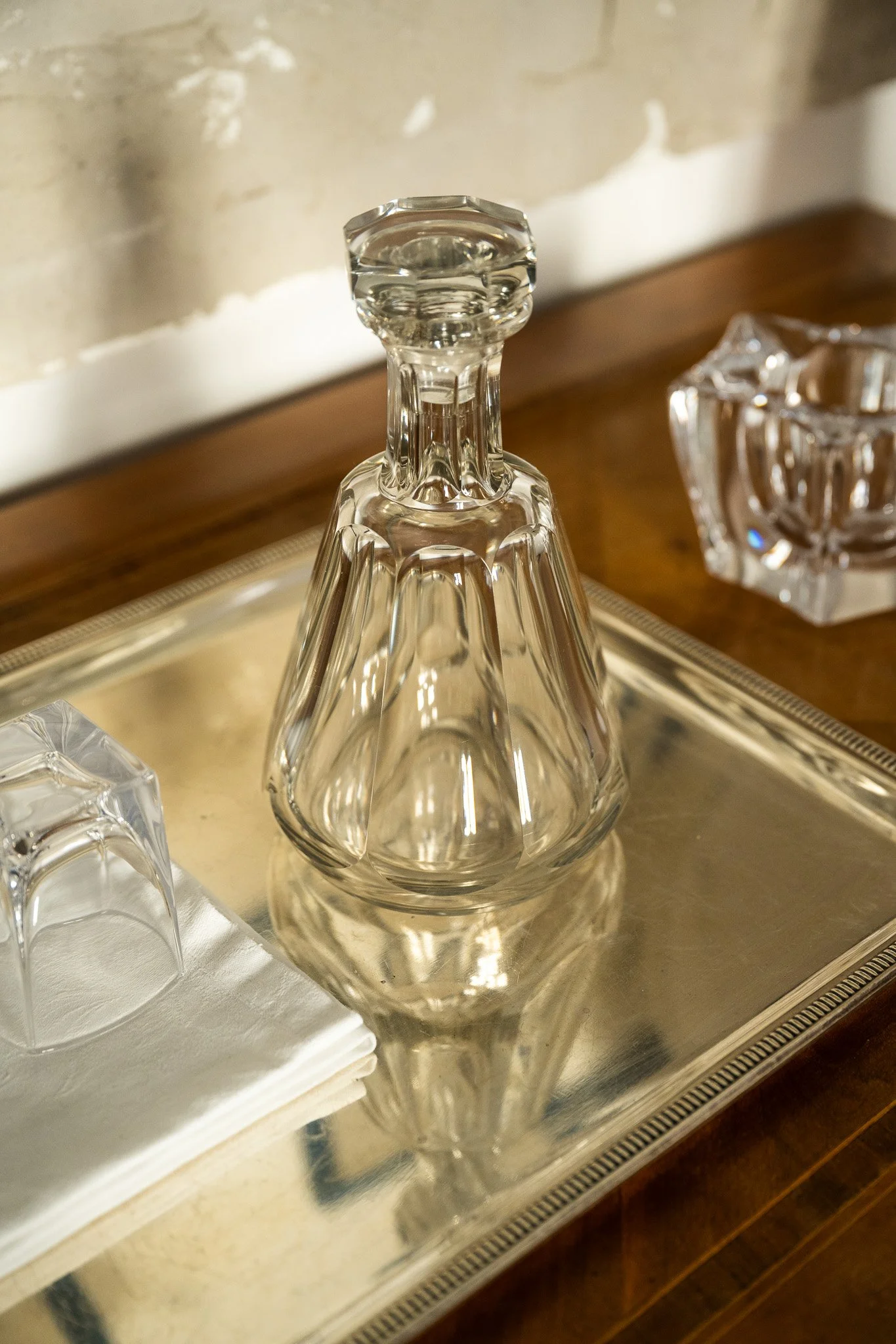 Une carafe en cristal vide avec un bouchon, placée sur un plateau en métal miroir, avec des verres en cristal à côté et des serviettes blanches en tissu, dans un décor intérieur élégant.