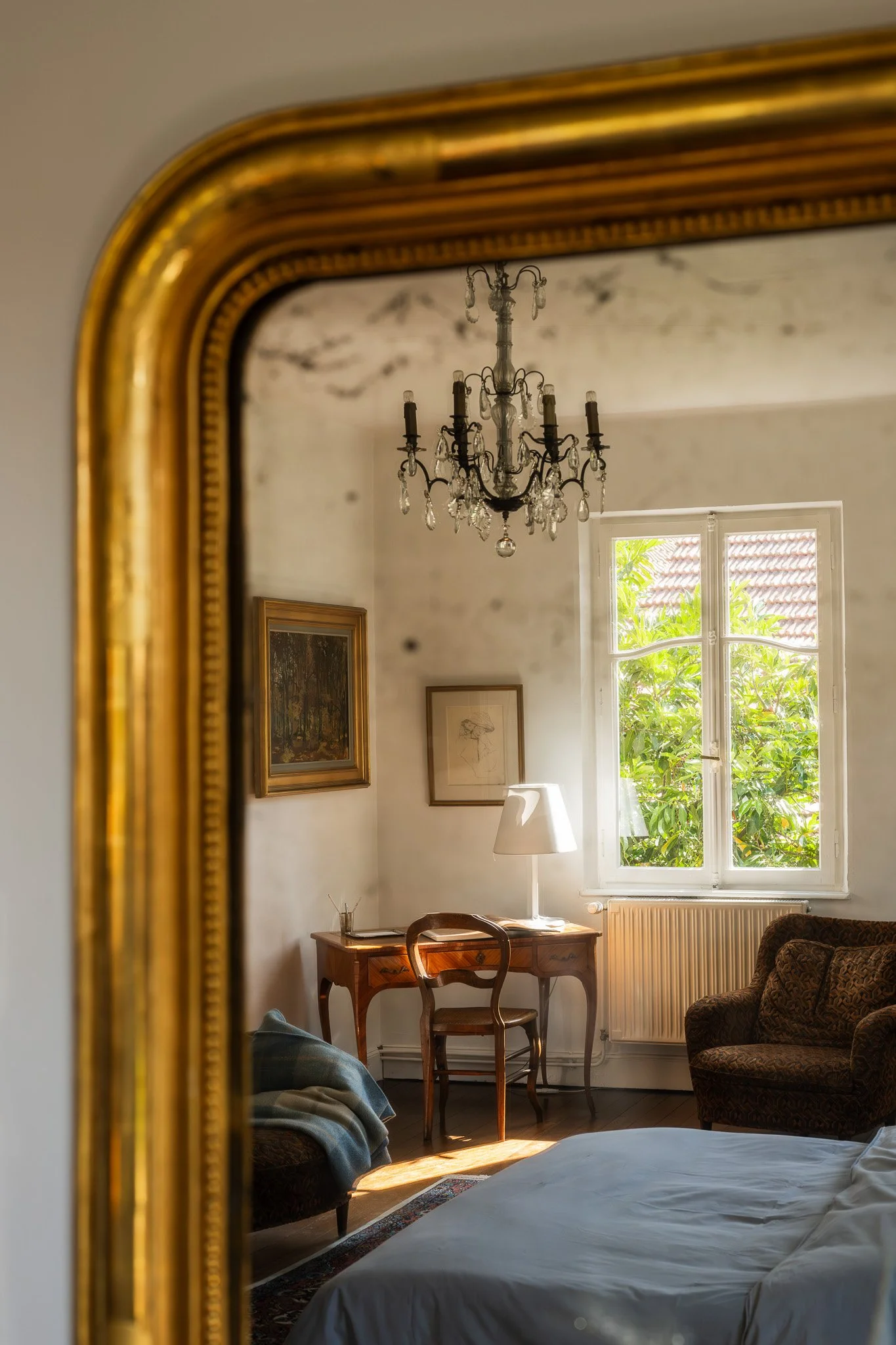 Vue d'une chambre à travers un miroir doré, montrant un lit, un fauteuil, une commode avec une lampe et des tableaux au mur, une fenêtre laissant entrer la lumière naturelle et des plantes à l'extérieur.