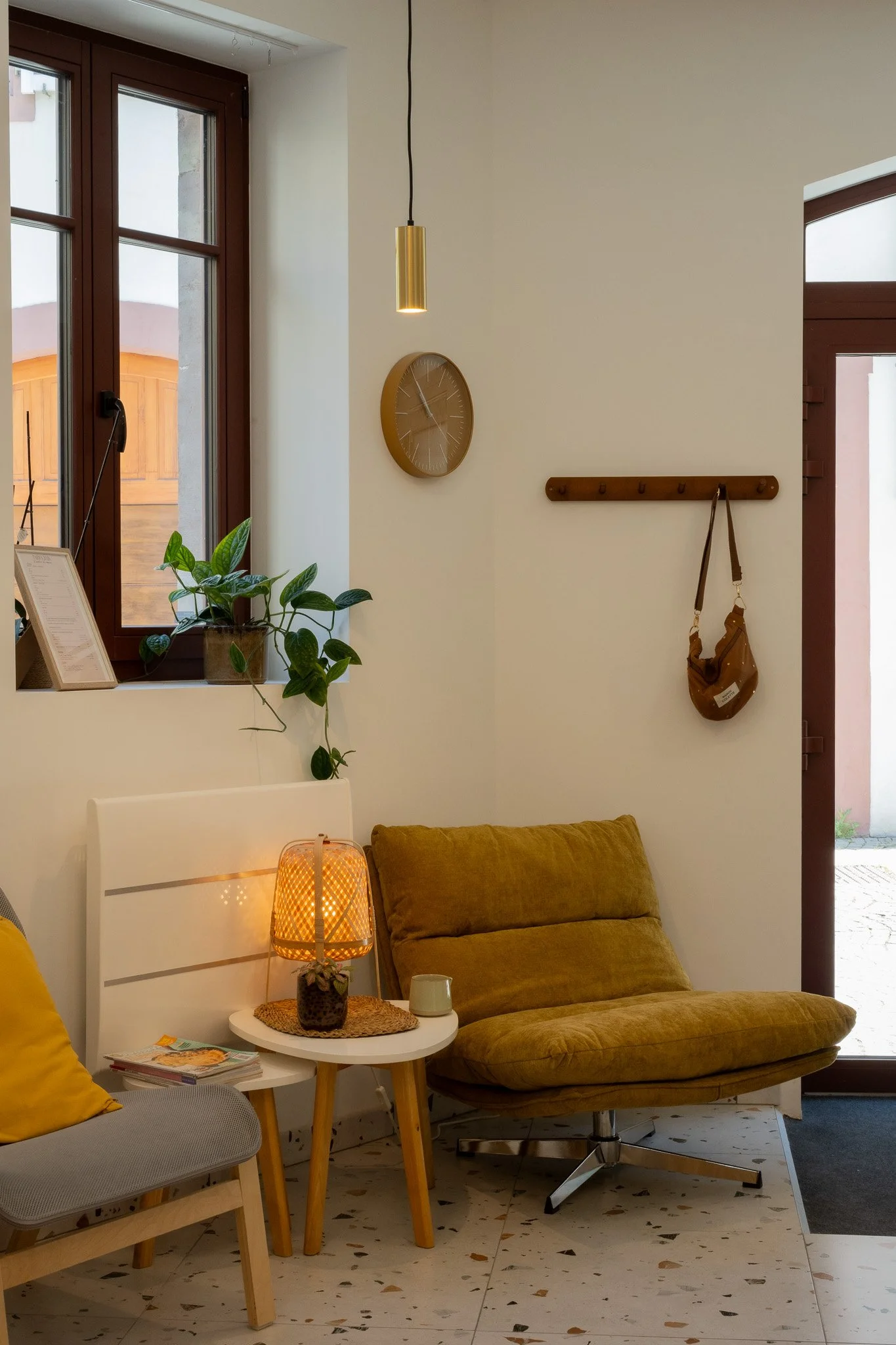 Espace attente d'un institut de beauté avec fauteuils jaunes, une lampe en osier, une table blanche avec des magazines, un mur blanc avec une horloge et un porte-manteaux en bois avec un sac en bandoulière.