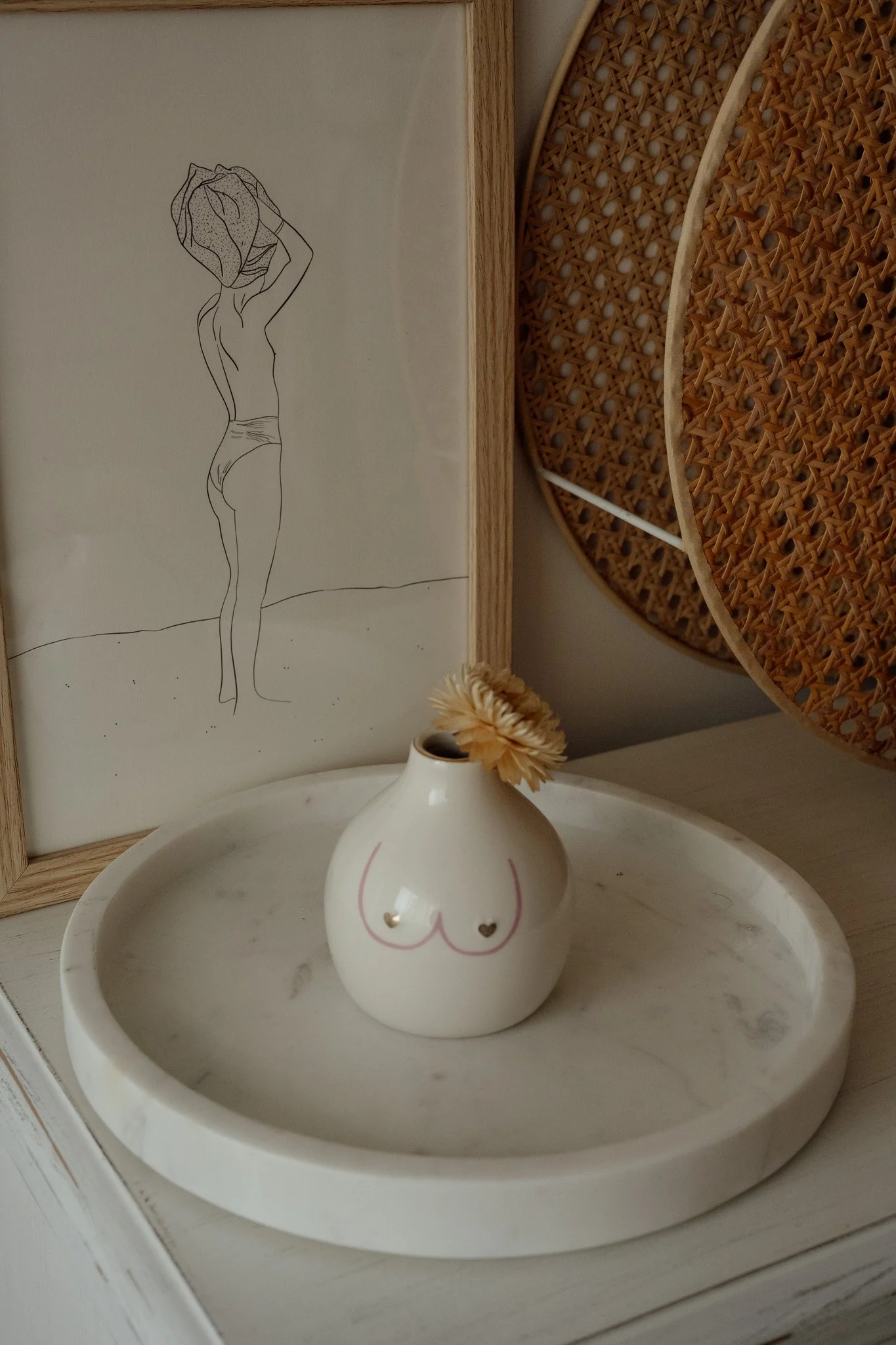 Une illustration minimaliste d'une femme en sous-vêtements, de dos, tenant un sac, dessin noir sur fond blanc, en arrière-plan décor de style naturel avec paniers en rotin et une plante