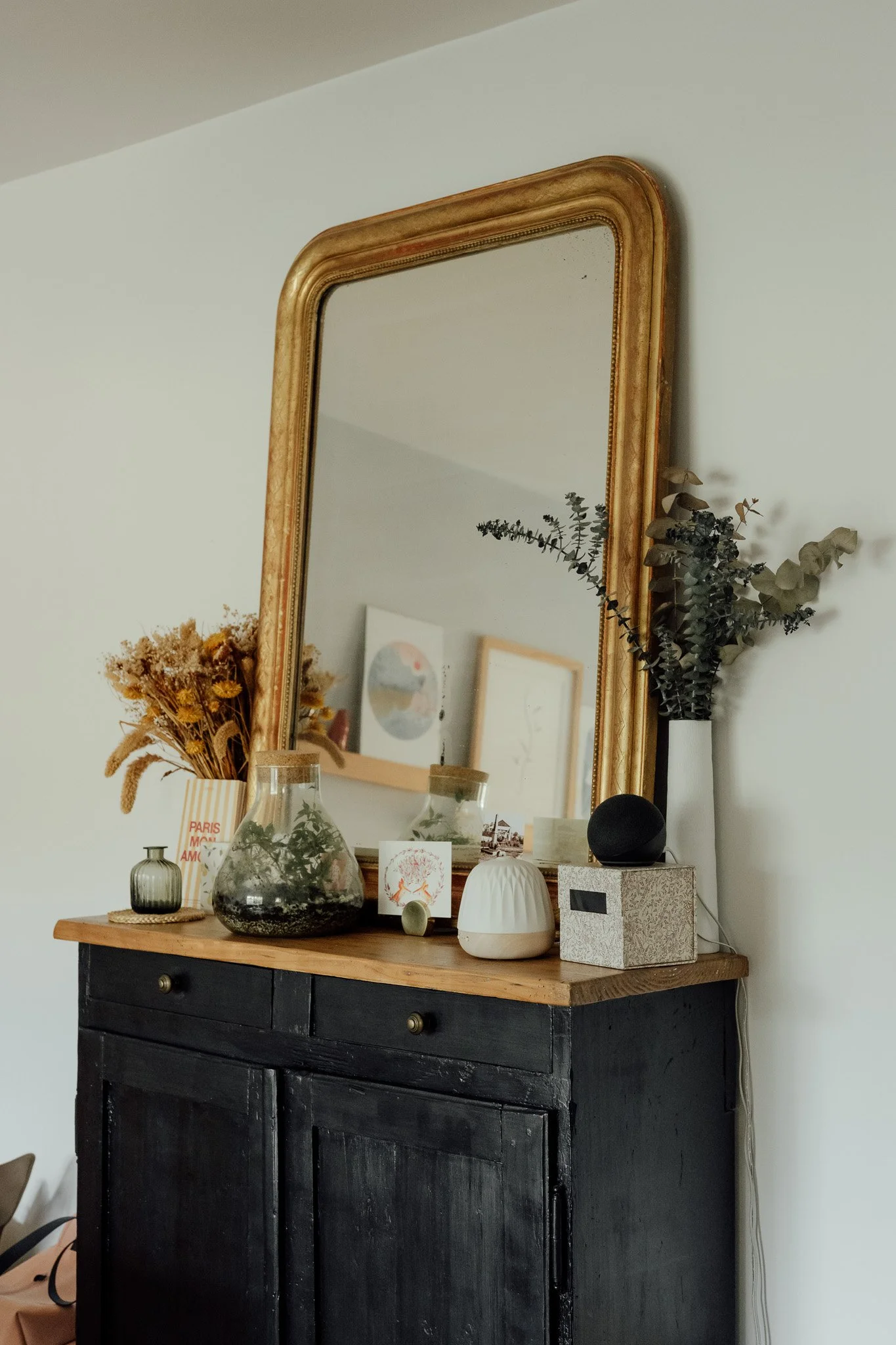 Un miroir vieux de style antique accroché à un mur blanc, avec des décorations de plantes séchées, vases, et accessoires sur une console en bois noir en dessous.