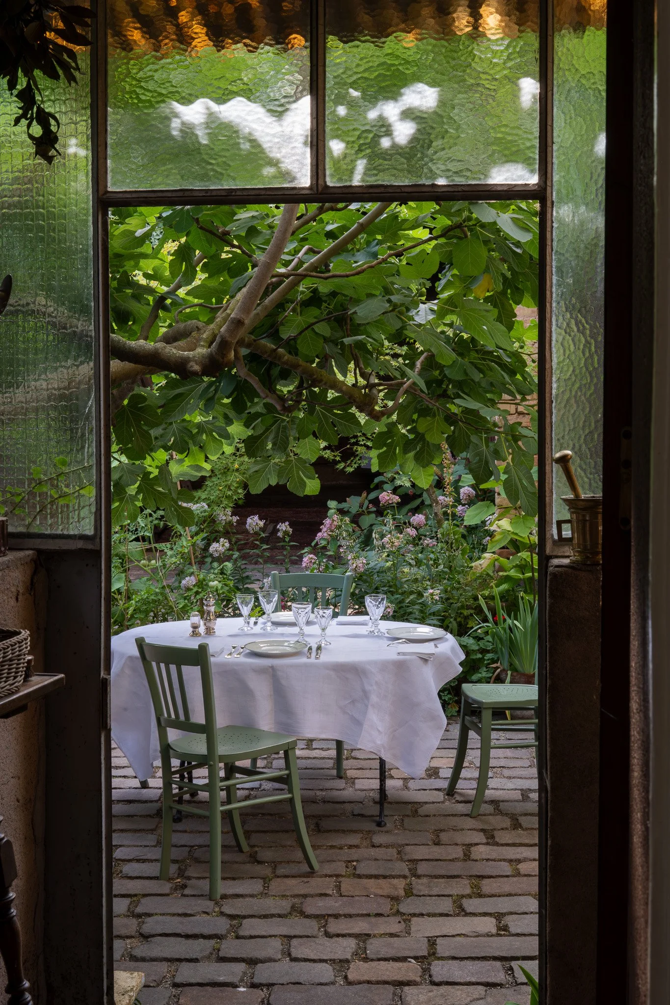 Une table dressée avec une nappe blanche, des verres et des assiettes, située dans un patio avec des chaises vertes, entouré de plantes et d'arbres, vu à travers une porte-fenêtre en verre.