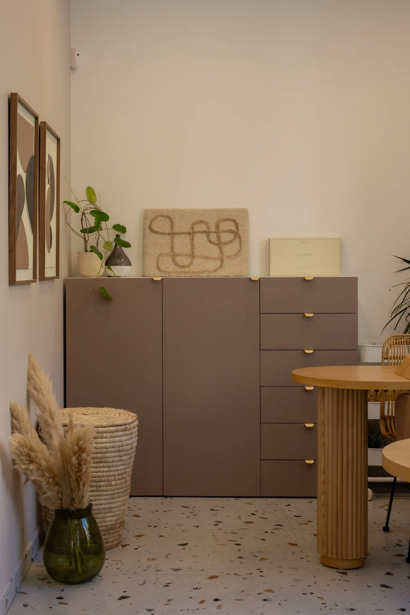 Vue intérieure d'un institut de beauté avec un meuble gris, une plante en pot, œuvres d'art abstrait, une boîte rectangulaire, un tapis en fibre naturelle et un vase en verre vert au sol.
