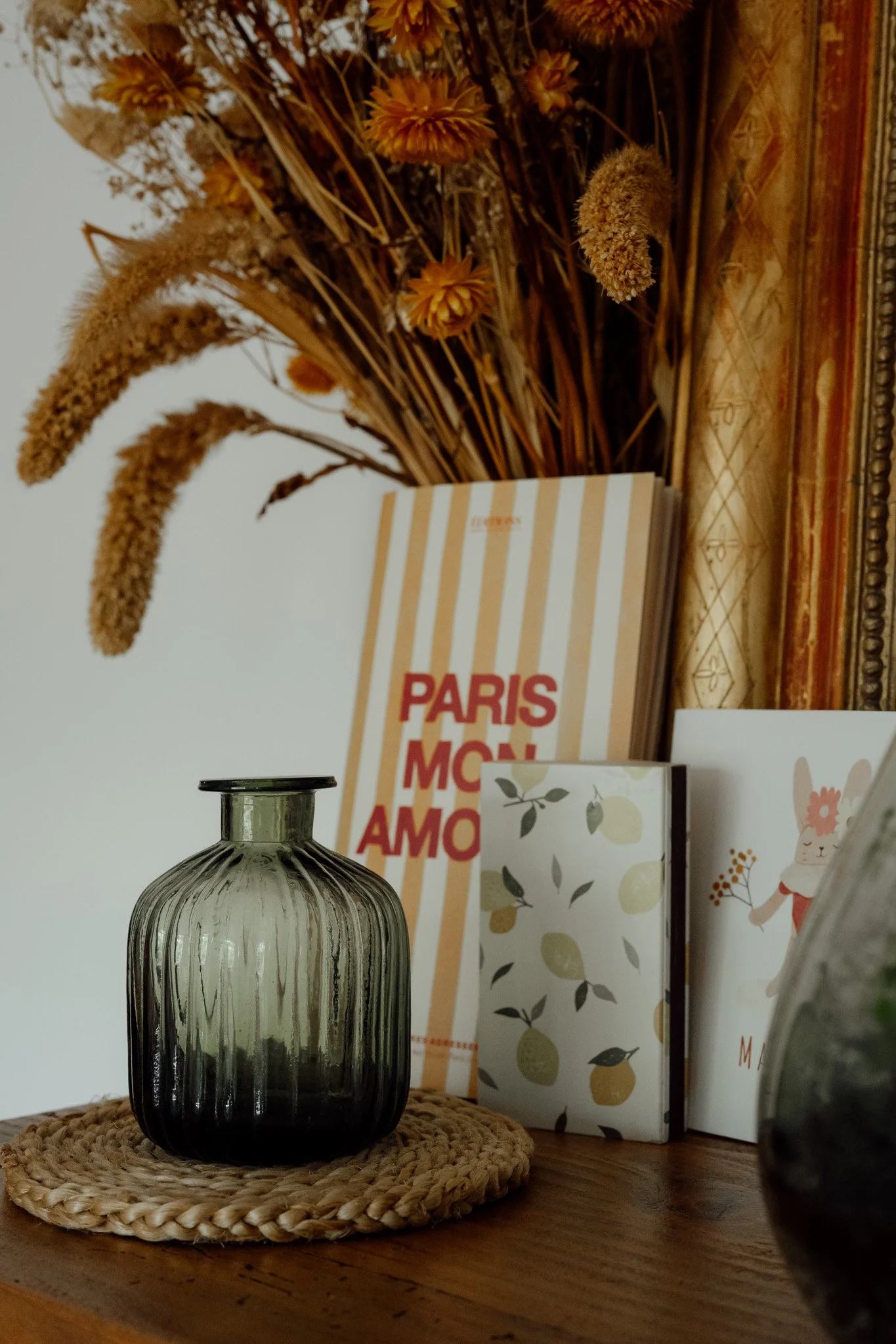 Un vase en verre gris foncé avec des rayures verticales, placé sur un support en bois, en face d'un livre avec une couverture rayée beige et blanche, et d'autres livres avec des illustrations de citrons et de lapins, tout cela sur une table en bois. 