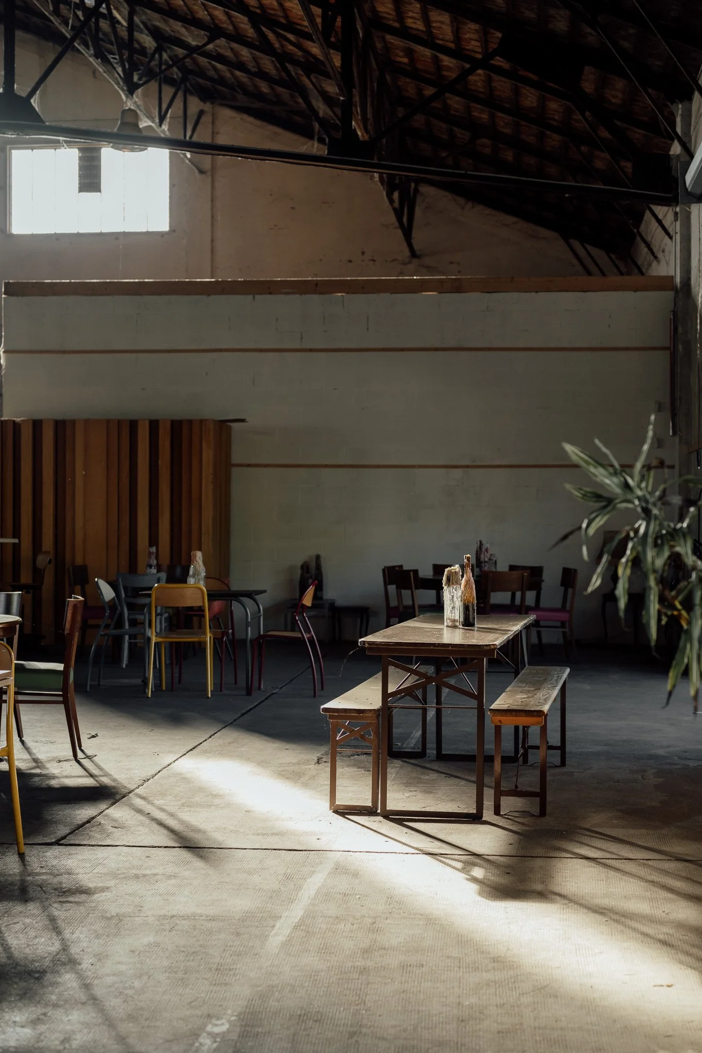 Une salle avec des tables en bois et des chaises, éclairée par la lumière du soleil passant par une fenêtre en haut, dans un espace de style industriel avec plafond en bois et murs bruts.