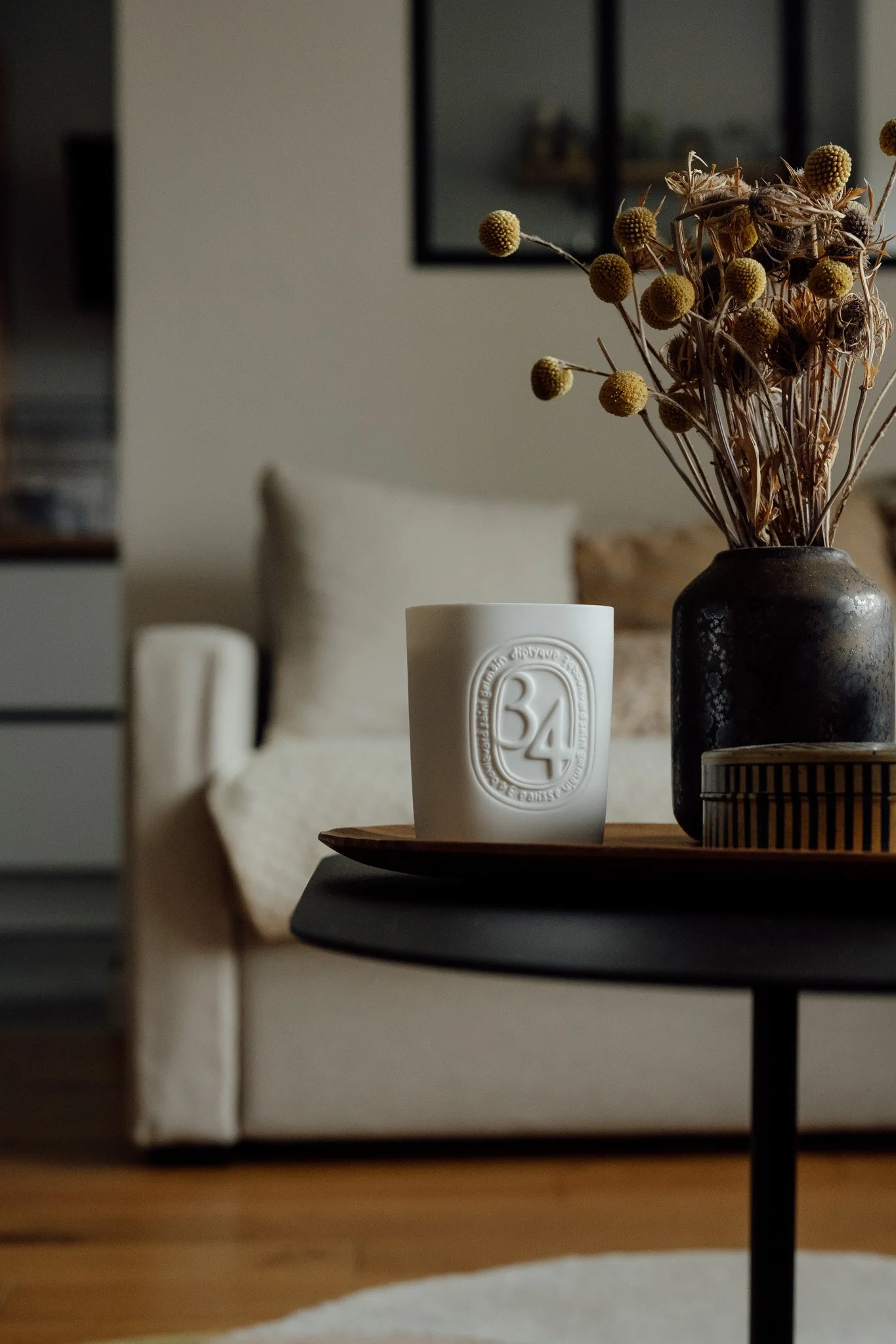 Une lampe blanche avec le chiffre 34 en relief, située sur une table en bois, à côté d'un vase noir contenant des branches sèches avec des fleurs séchées.