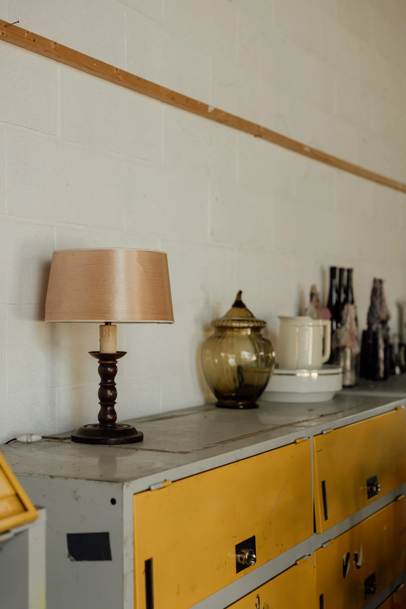  Lampe de table en bois avec abat-jour en tissu beige, décorant un meuble ancien avec une surface en marbre, entouré de divers objets en verre et céramique, sur un mur en briques blanches 
