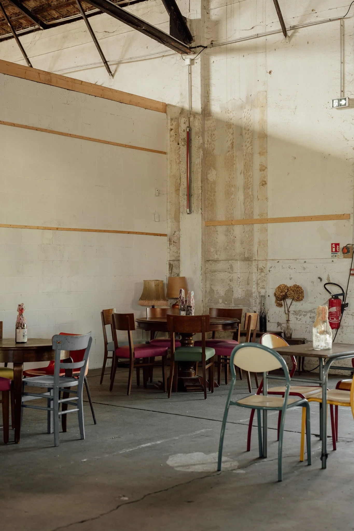 Intérieur d'un espace de restauration ou cafétéria avec plusieurs tables et chaises en bois et en métal, murs en pierre peinte, sol en béton, éclairage industriel, décor simple avec plantes en vase, et équipement de sécurité comme un extincteur.