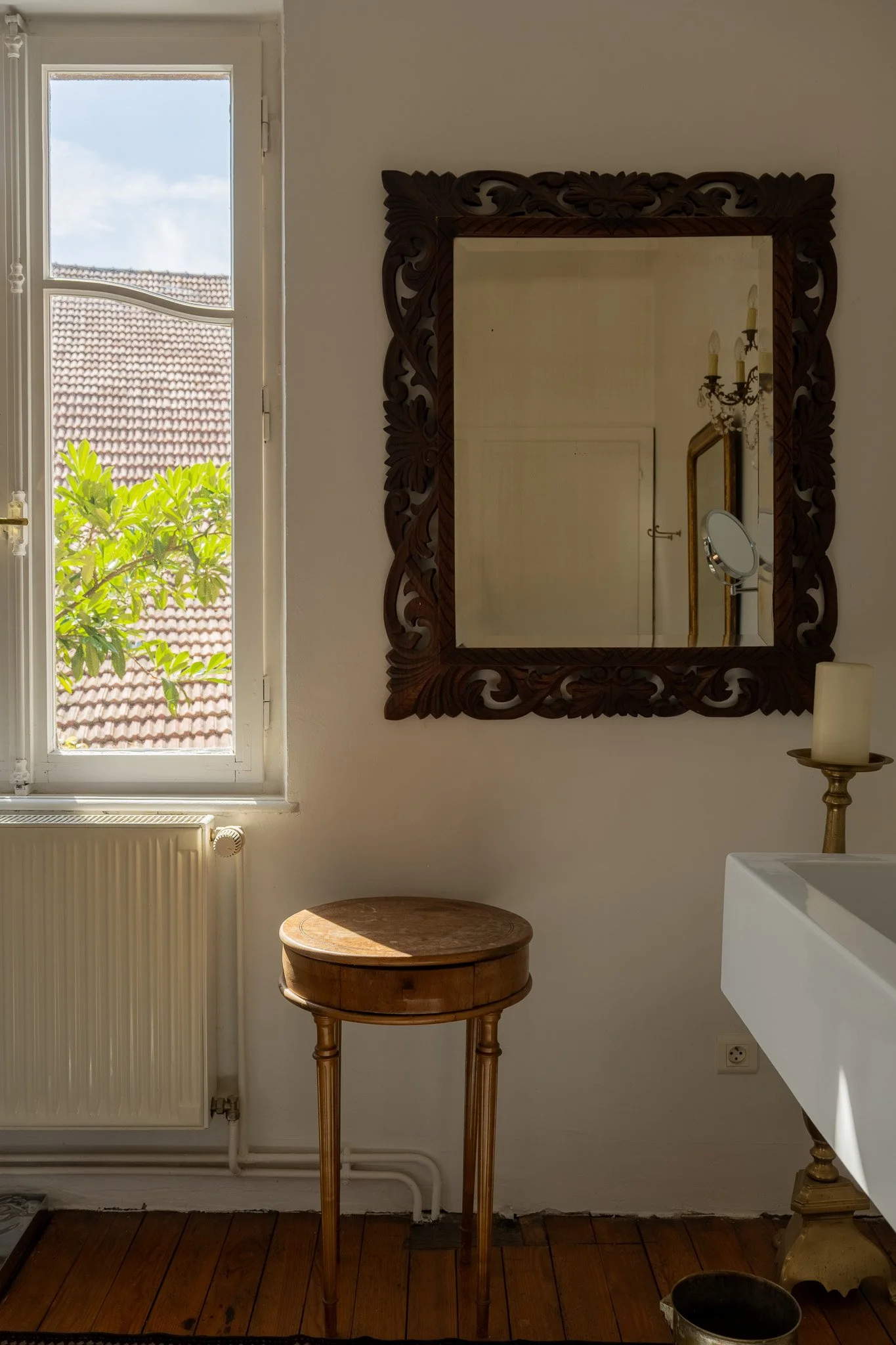 Intérieur d'une pièce avec une fenêtre laissant entrer la lumière, un miroir encadré de bois sculpté, une table en bois, une bougie sur un support doré, un radiateur blanc, et un sol en parquet en bois.