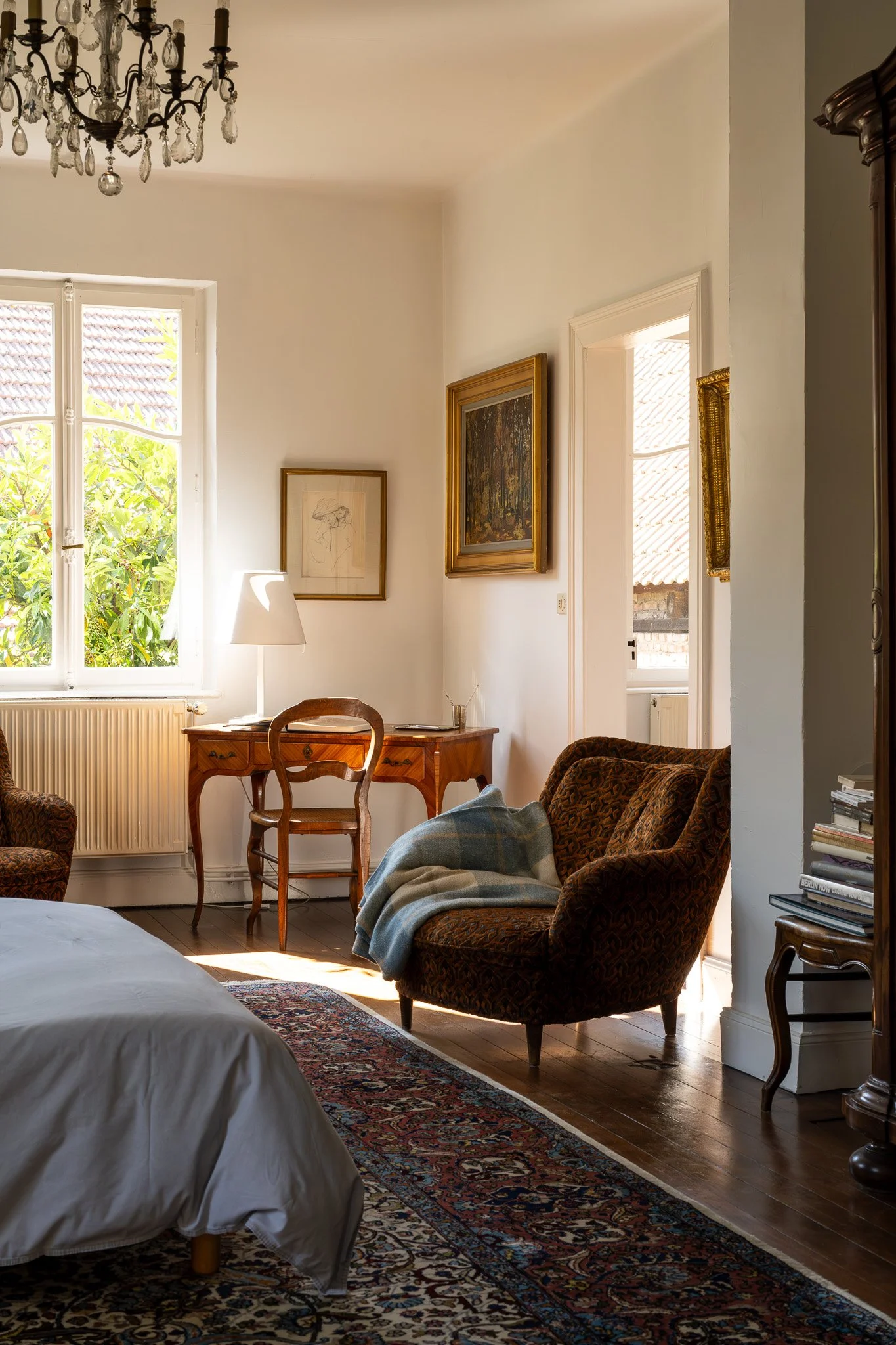 Un salon avec un canapé, une chaise en bois, des tableaux encadrés sur le mur, une lampe de chevet, une fenêtre avec des volets laissant entrer la lumière, et un tapis oriental.