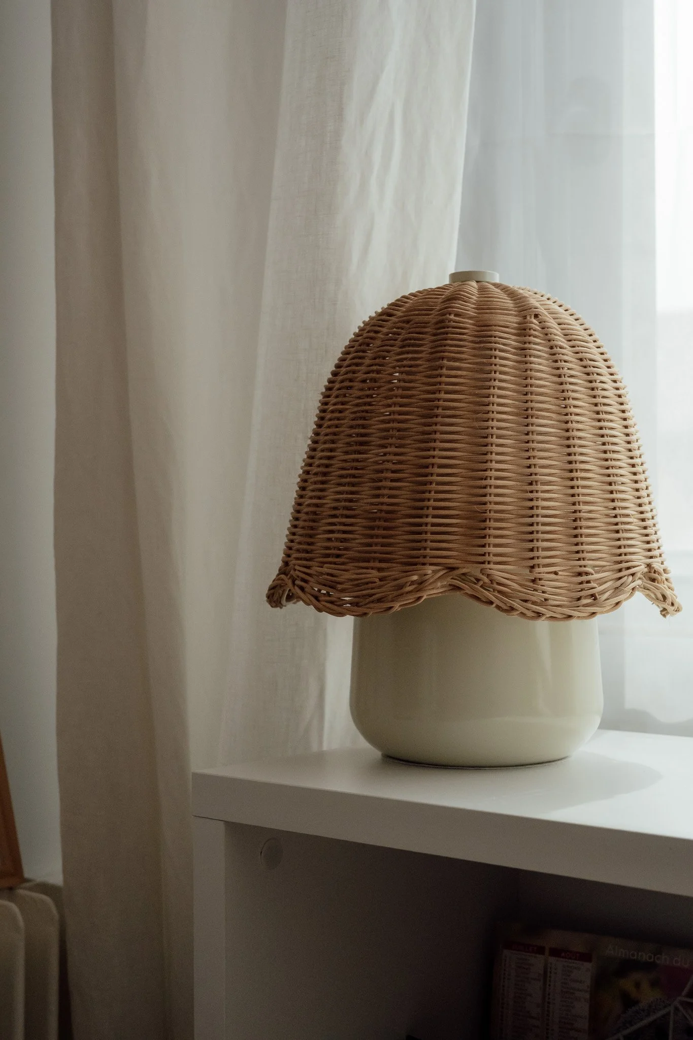 Une lampe en rotin posée sur une étagère blanche, avec un conteur blanc en dessous, dans une pièce lumineuse avec rideaux blancs en arrière-plan.