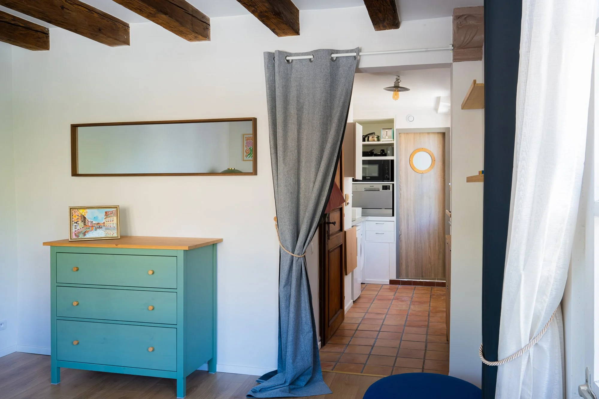 Intérieur d'un appartement avec un meuble bleu vif, un miroir allongé, un tableau devant, une kitchenette avec micro-ondes, four et étagères, un mur blanc, un tapis en terre cuite au sol, porte d'entrée en bois, et un rideau blanc.