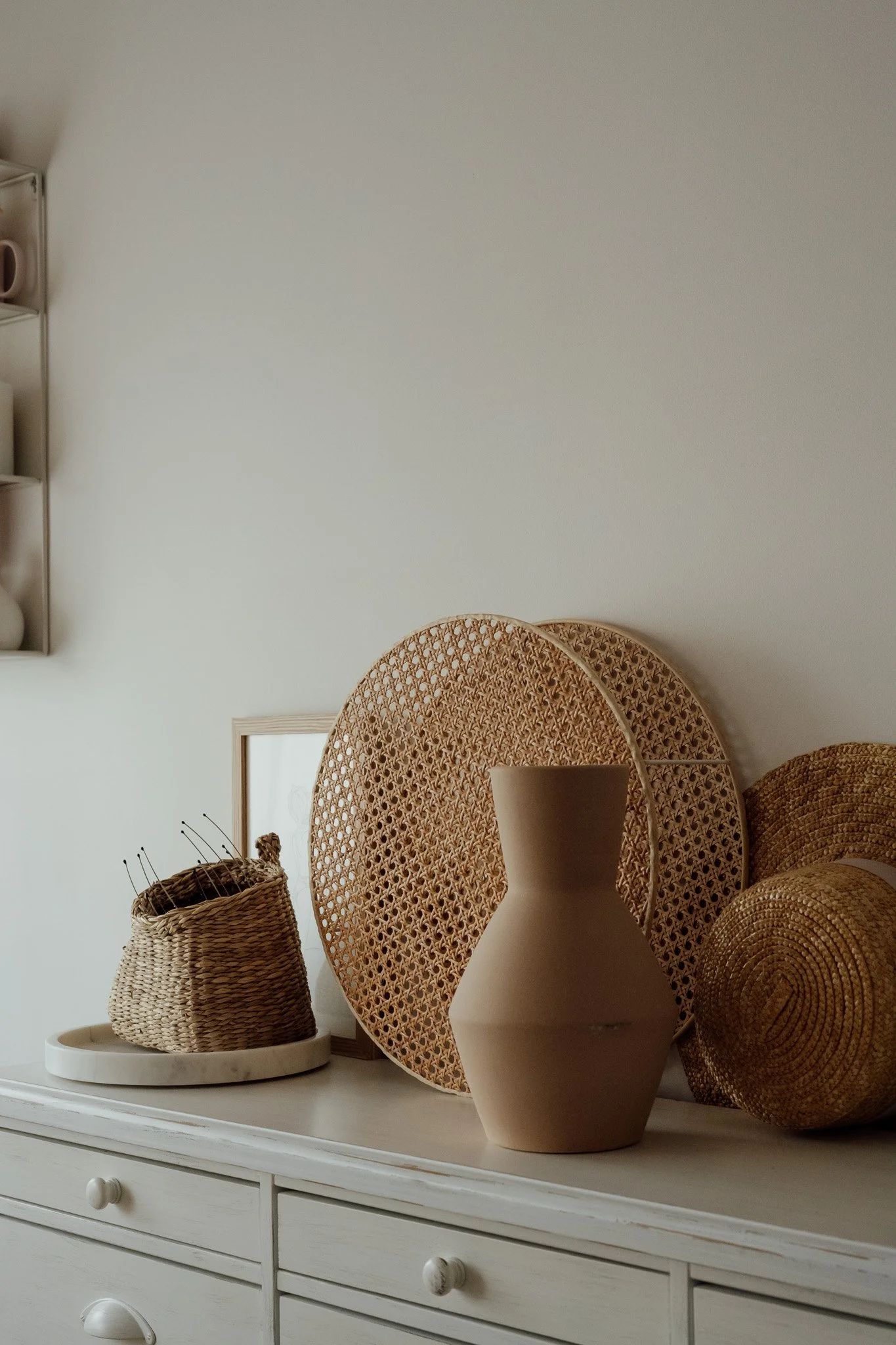 Une vitrine blanche contenant des paniers en osier, une grande jarre beige, des assiettes en bois et un miroir rond en rotin. Le tout décoratif et élégant, dans un style naturel et minimaliste.