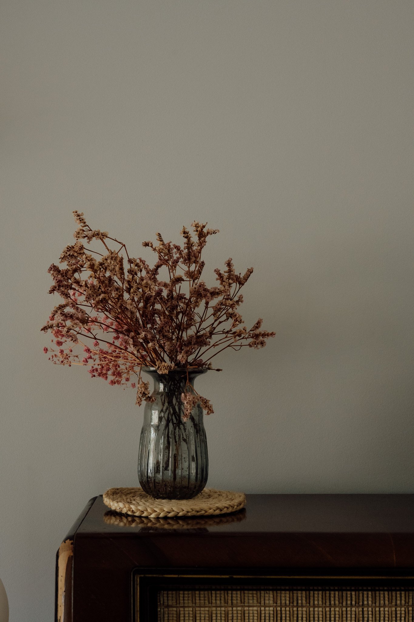 Un bouquet de fleurs séchées dans un vase en verre noir, placé sur une table en bois sombre, avec un mur gris en arrière-plan.
