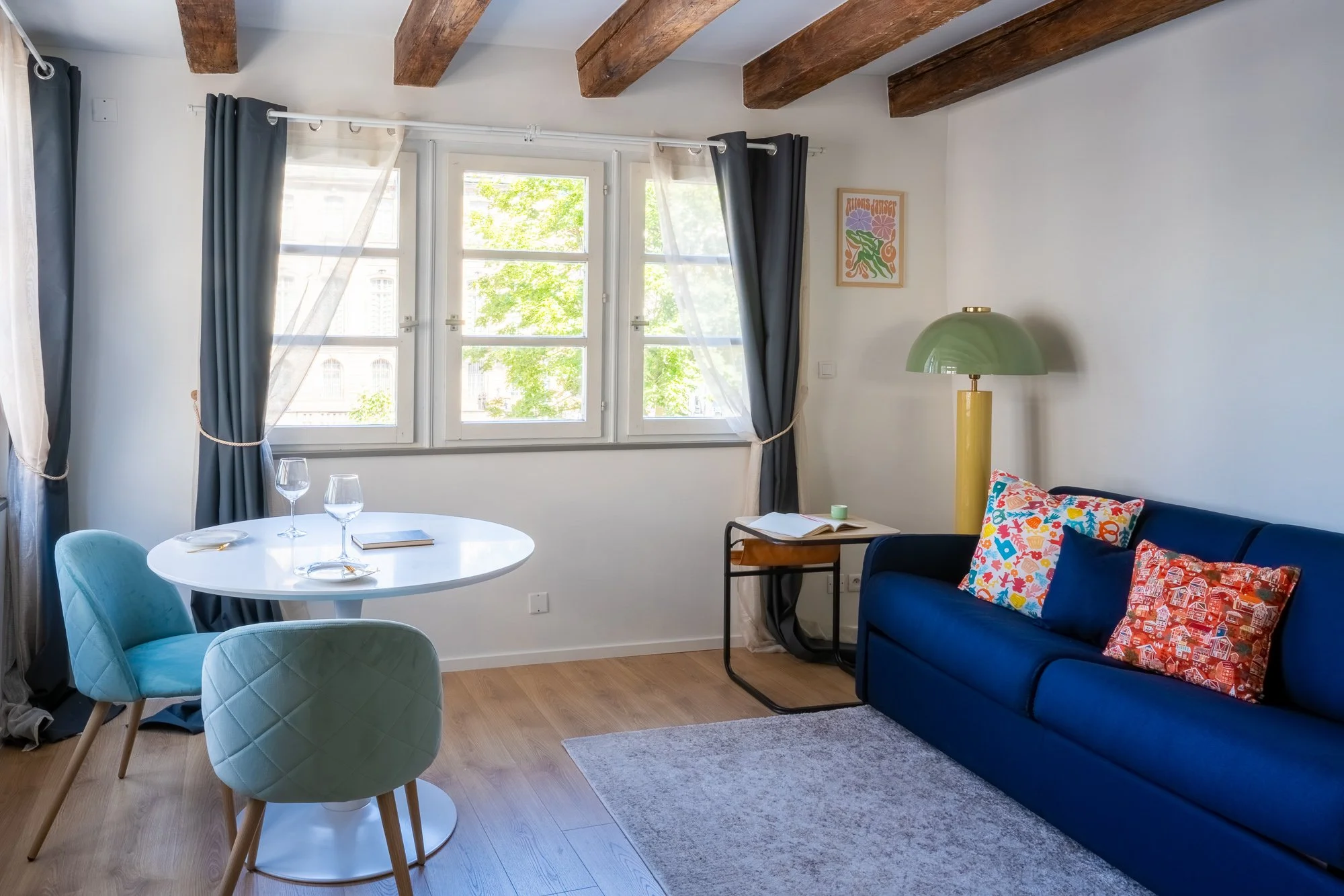 Salon de séjour avec un canapé bleu, des coussins colorés, une table ronde blanche avec des verres et un livre, fenêtres avec rideaux gris et blancs, lampe verte sur pied, tableau coloré, parquet en bois, et un bureau avec un livre ouvert.
