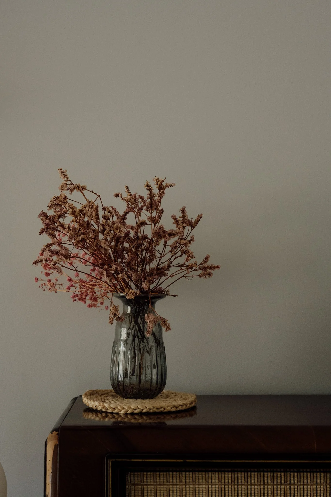 Un vase en verre transparent contenant des branches de fleurs séchées, placé sur une petite table noire avec un tapis en rotin dessous, contre un mur gris.
