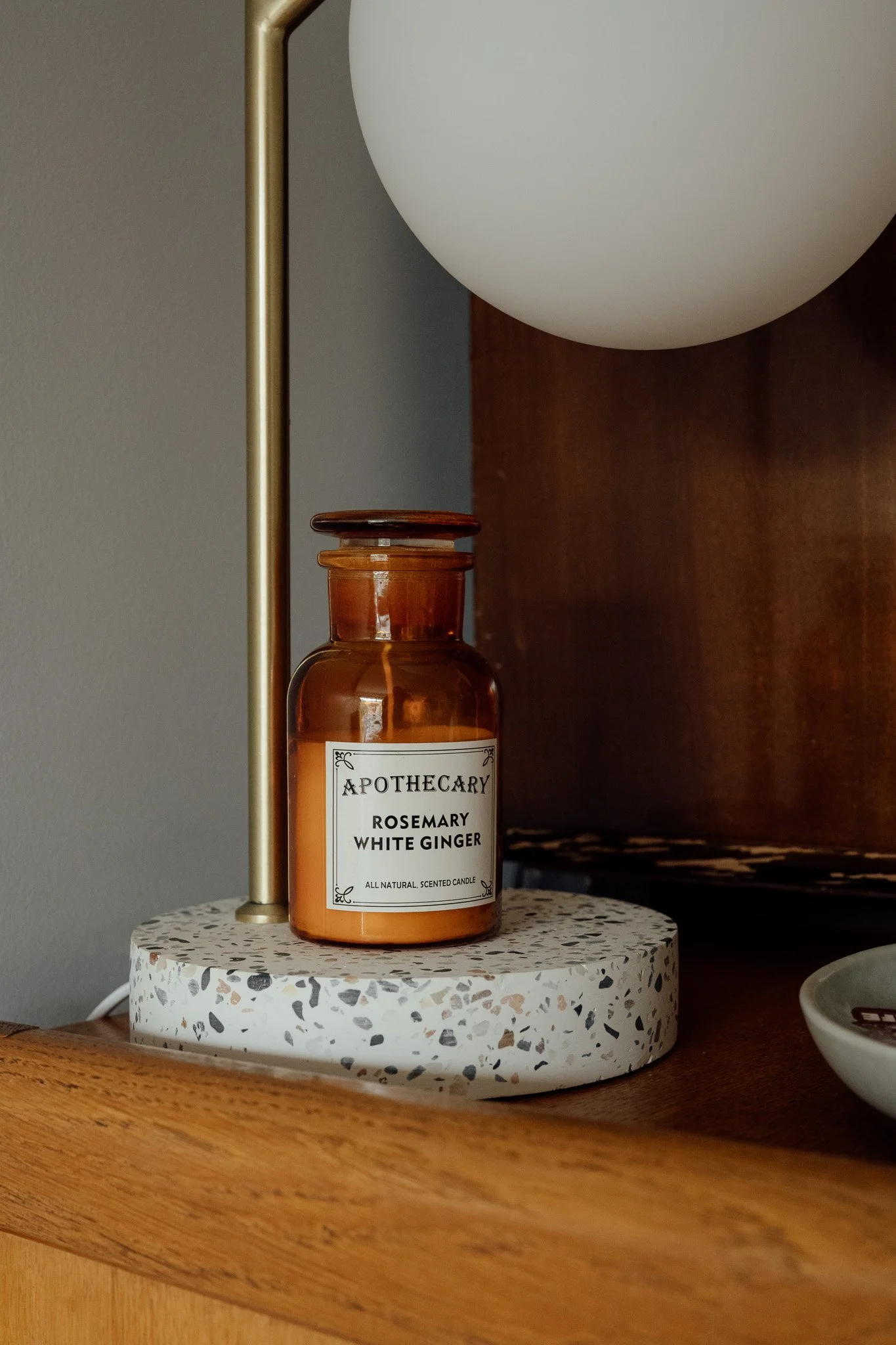Une bougie aromatique dans un pot en verre marron, étiquetée « Apothecary Rosemary White Ginger ». La bougie est placée sur une surface en terrazzo avec une lampe à côté.