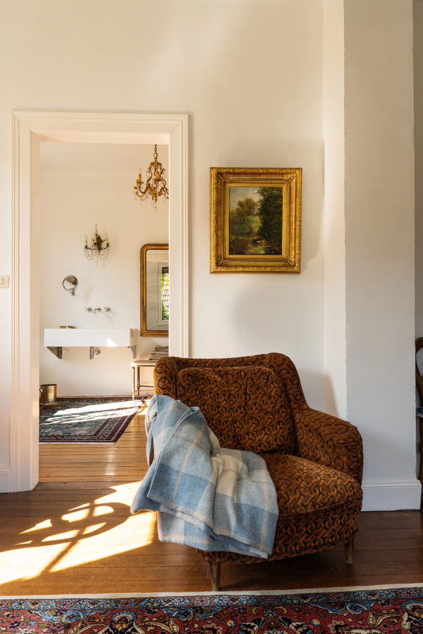 Vue intérieure d'un salon avec un canapé aux motifs géométriques, un plaid à carreaux, un tapis persan, un miroir doré, un tableau représentant un paysage, et une porte menant à une salle de bain avec un chandelier en cristal