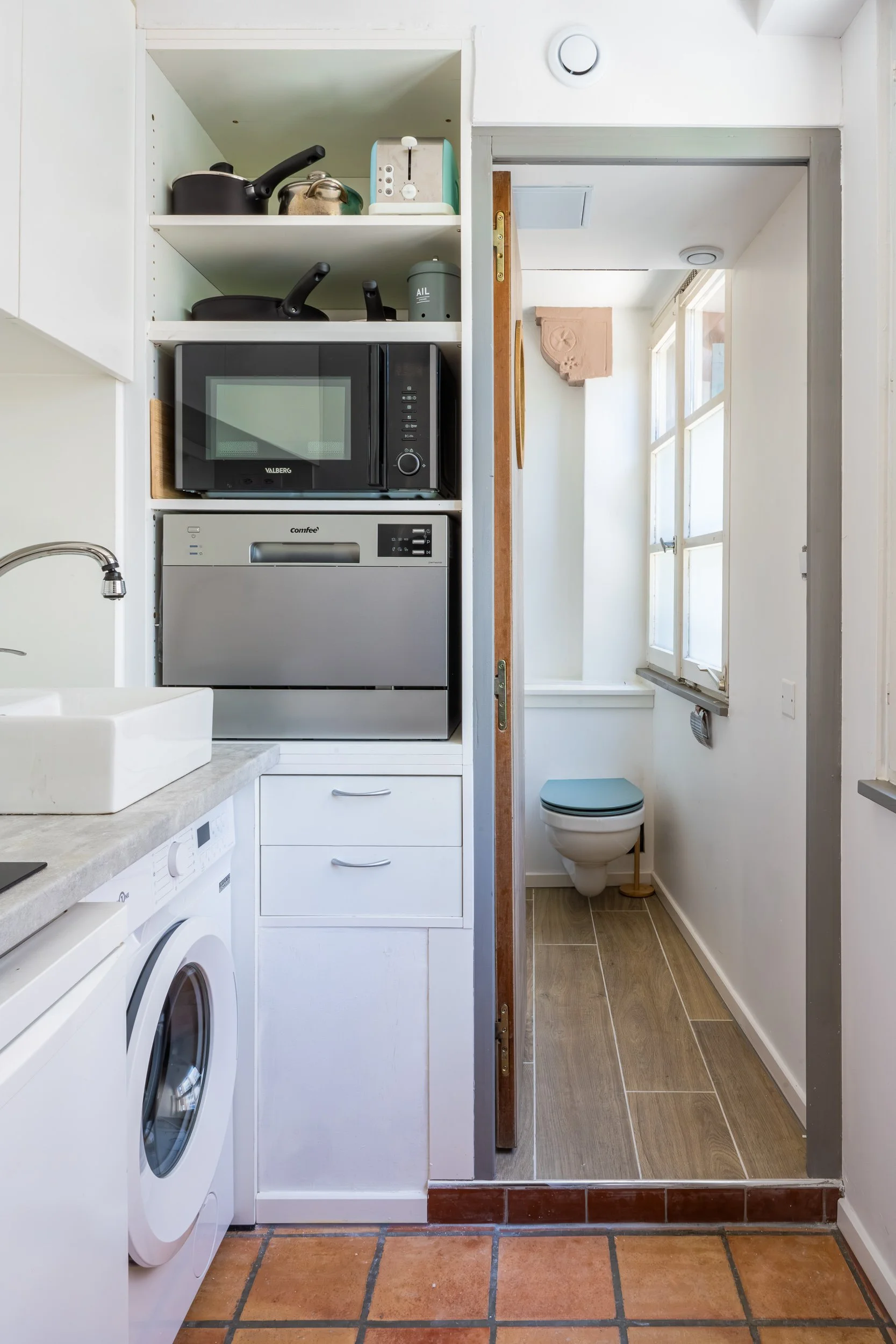 Coin buanderie avec lave-linge, sèche-linge, étagères contenant des casseroles, micro-ondes, et un tiroir à linge, séparée d'une salle de bain avec toilettes par une porte en bois.