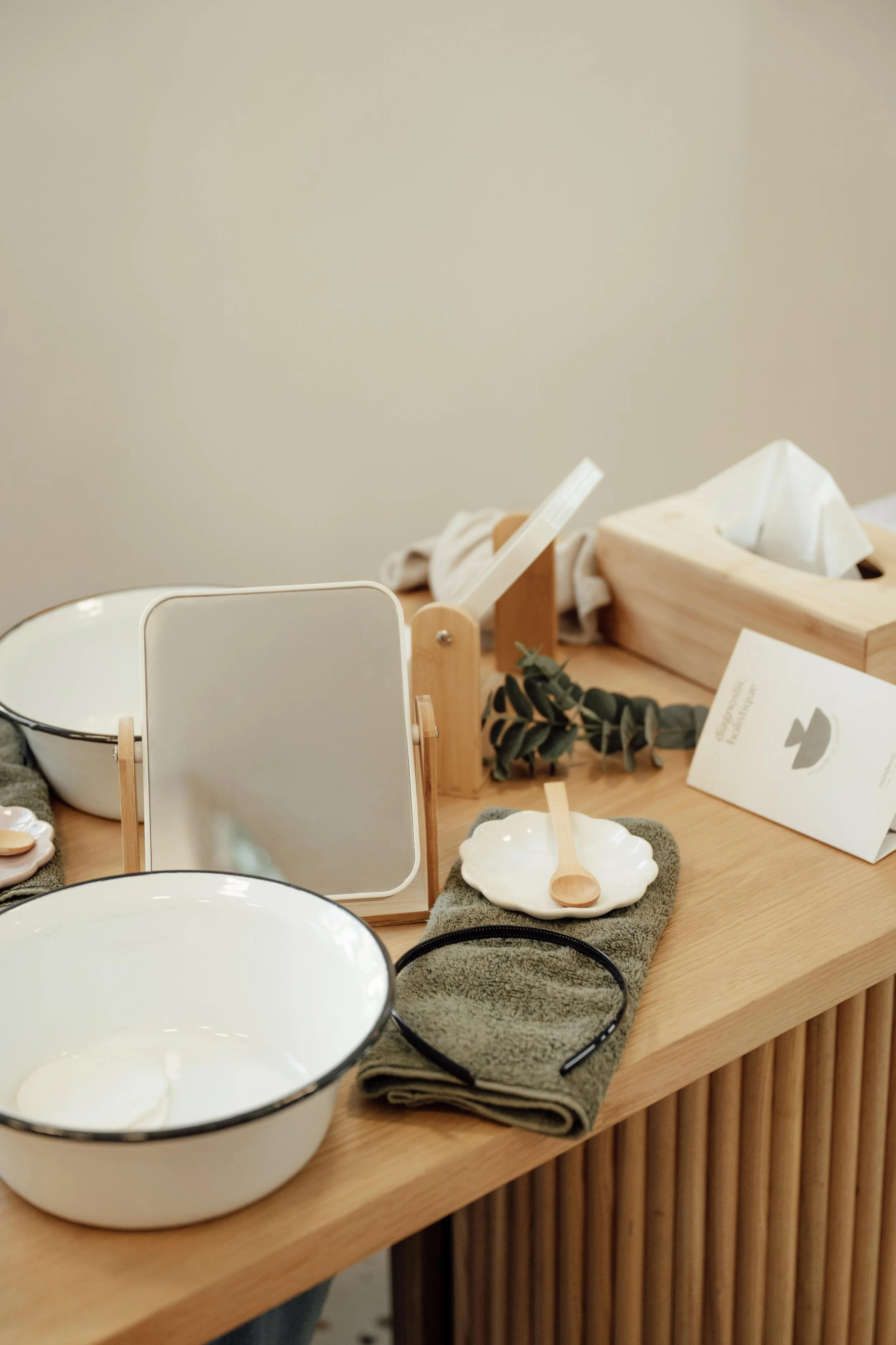 Vitrine de produits de soin à base de lin, y compris des bols en céramique, un miroir, une serviette en tissu, un pinceau en bois, un bandeau en tissu et une plante verte, présentation dans un décor minimaliste.