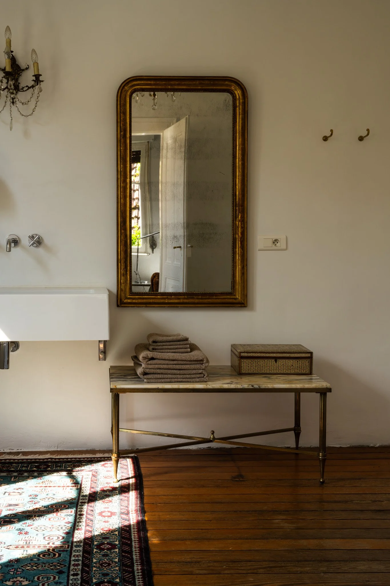 Un miroir ancien avec un cadre doré, un chandelier à gauche, une table en marbre avec des serviettes pliées, un petit coffre en bois, un tapis oriental, une pièce lumineuse avec une fenêtre