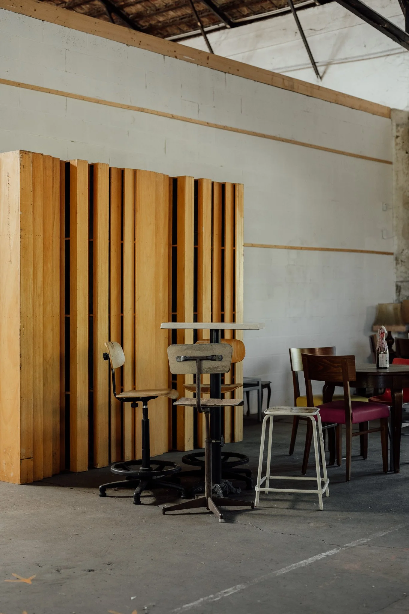 Intérieur industriel avec mobilier en bois, chaises de différentes tailles, tables, et cloison en bois, dans un espace avec un sol en béton