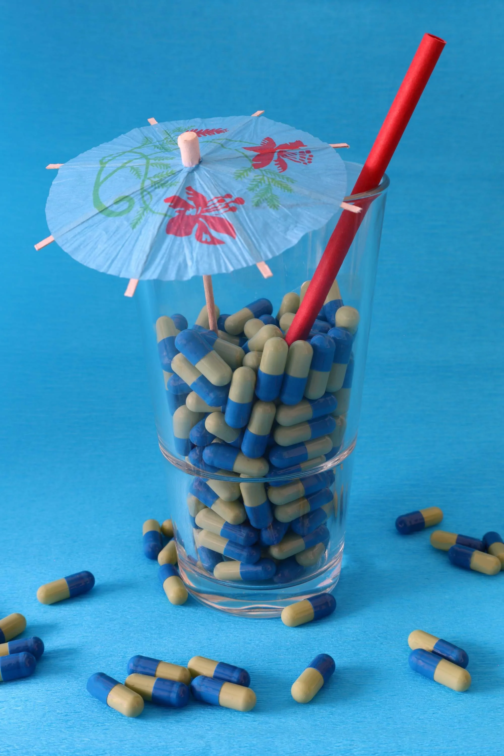 Ein Glas gefüllt mit Tabletten Strohhalm und Cocktailschirmchen vor blauem Hintergrund als Pillencocktail