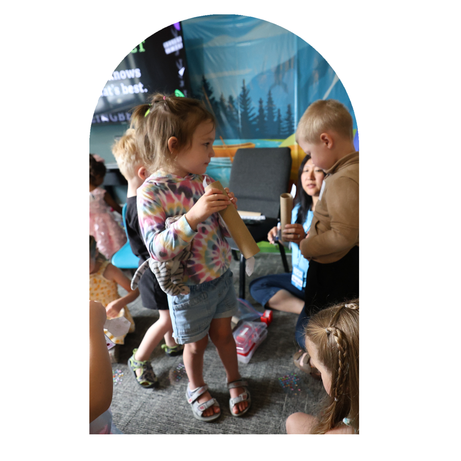 Children participating in a craft activity indoors, with a girl in a colorful tie-dye shirt and sandals holding a cardboard tube, and a boy in a tan jacket also holding a cardboard tube, with other kids and an adult woman in the background.