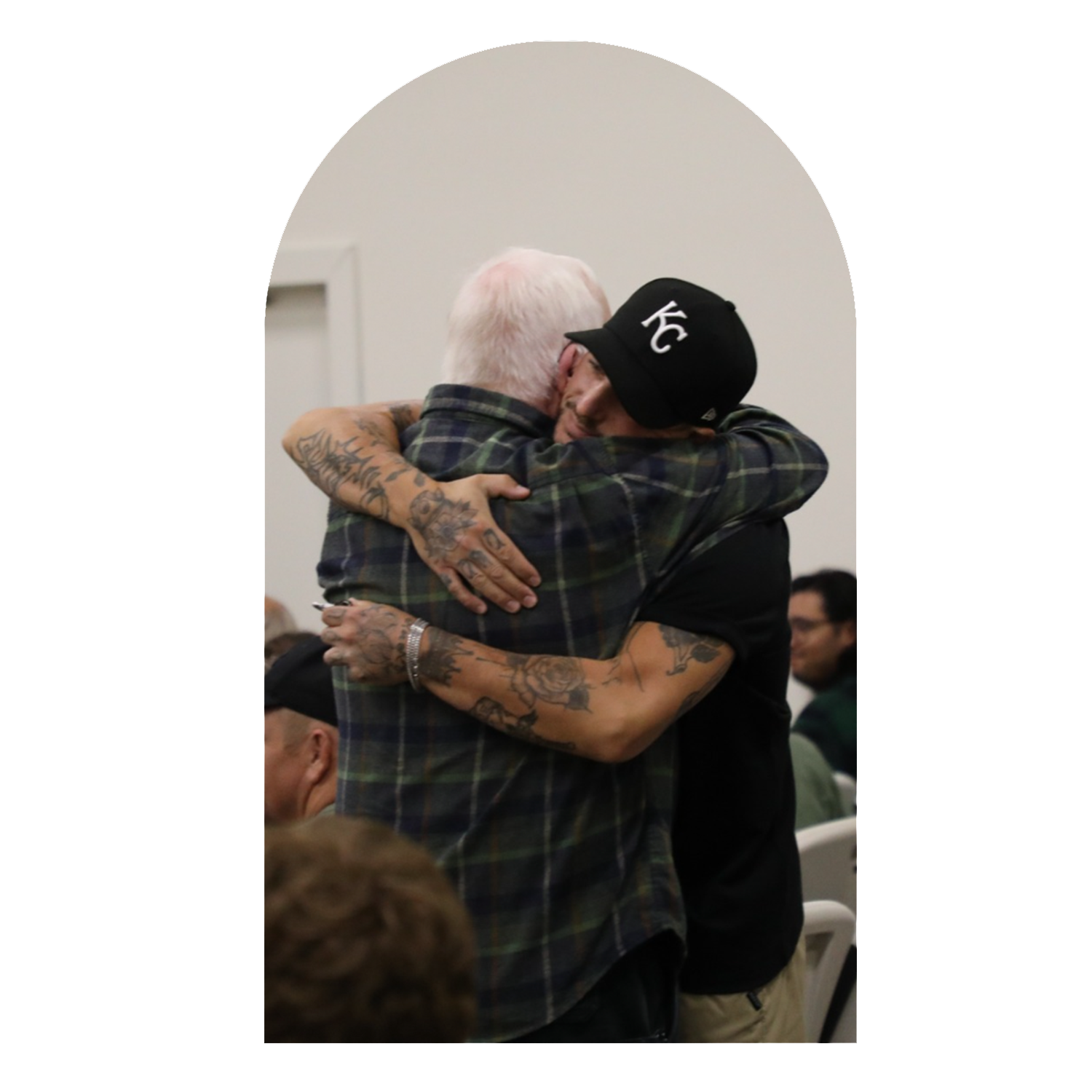 Two men hugging each other warmly, one with white hair and tattoos, the other wearing a black cap with 'KC' and glasses, in a group setting.
