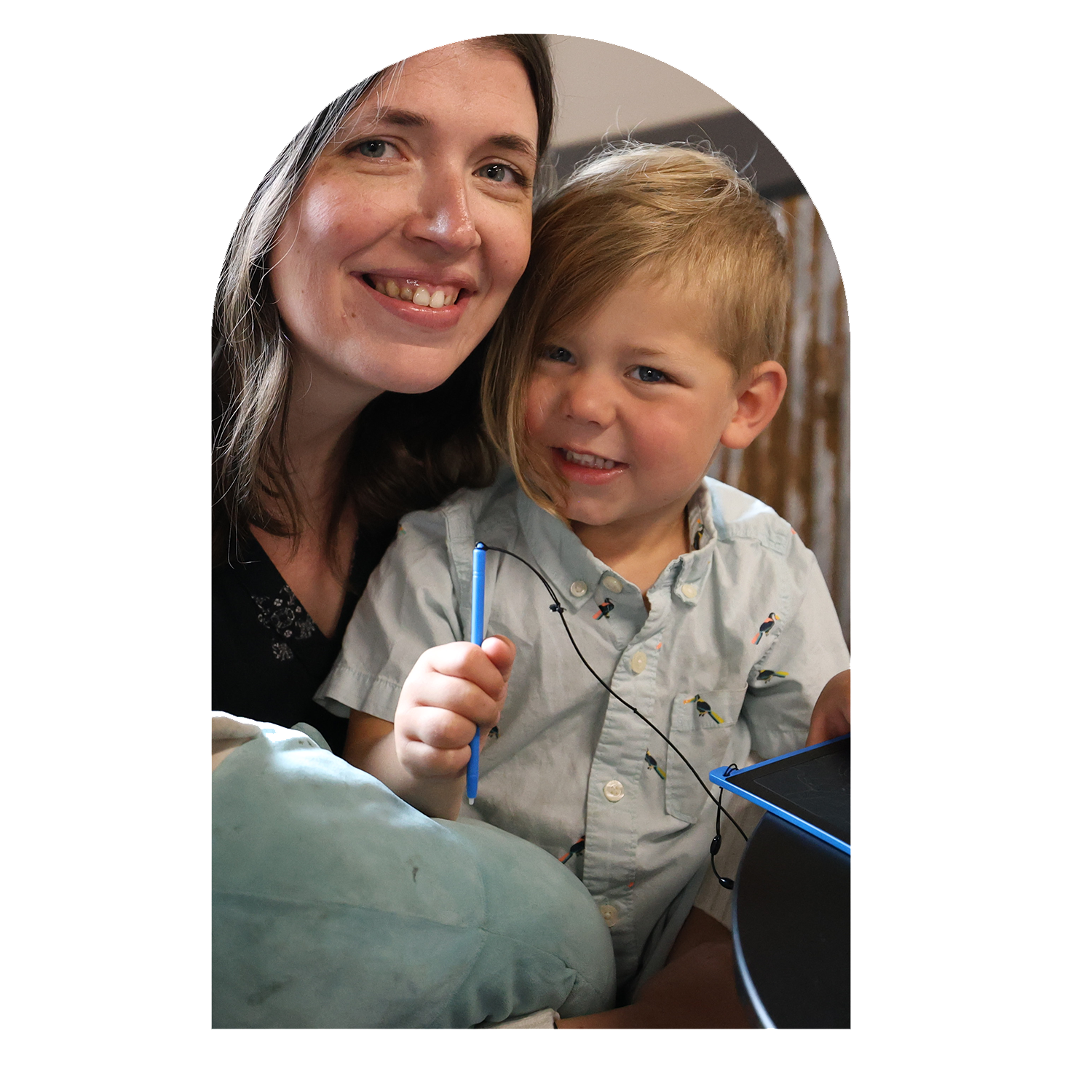 A woman and a young boy smiling together while playing with a tablet and a fishing game on a pen, indoors with a curtain in the background.