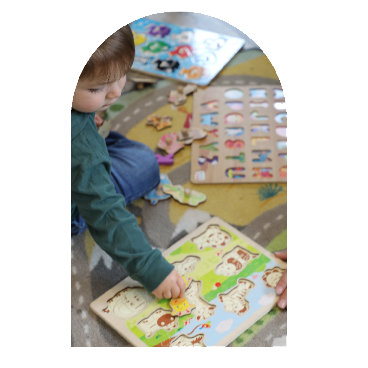 A young boy with brown hair sitting on a colorful patterned carpet, playing with wooden animal puzzles and a board with alphabet cutouts.