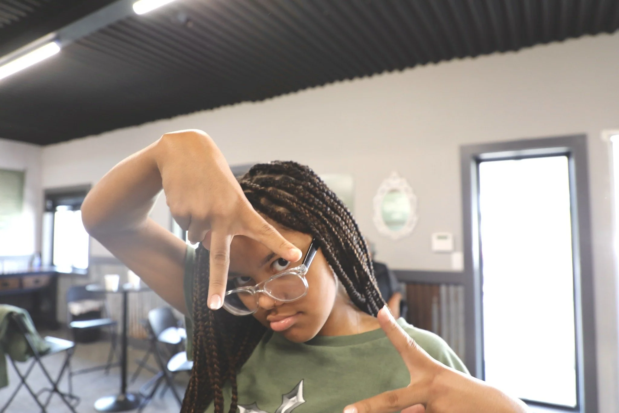 A young girl with braided hair and glasses making a playful pose indoors.