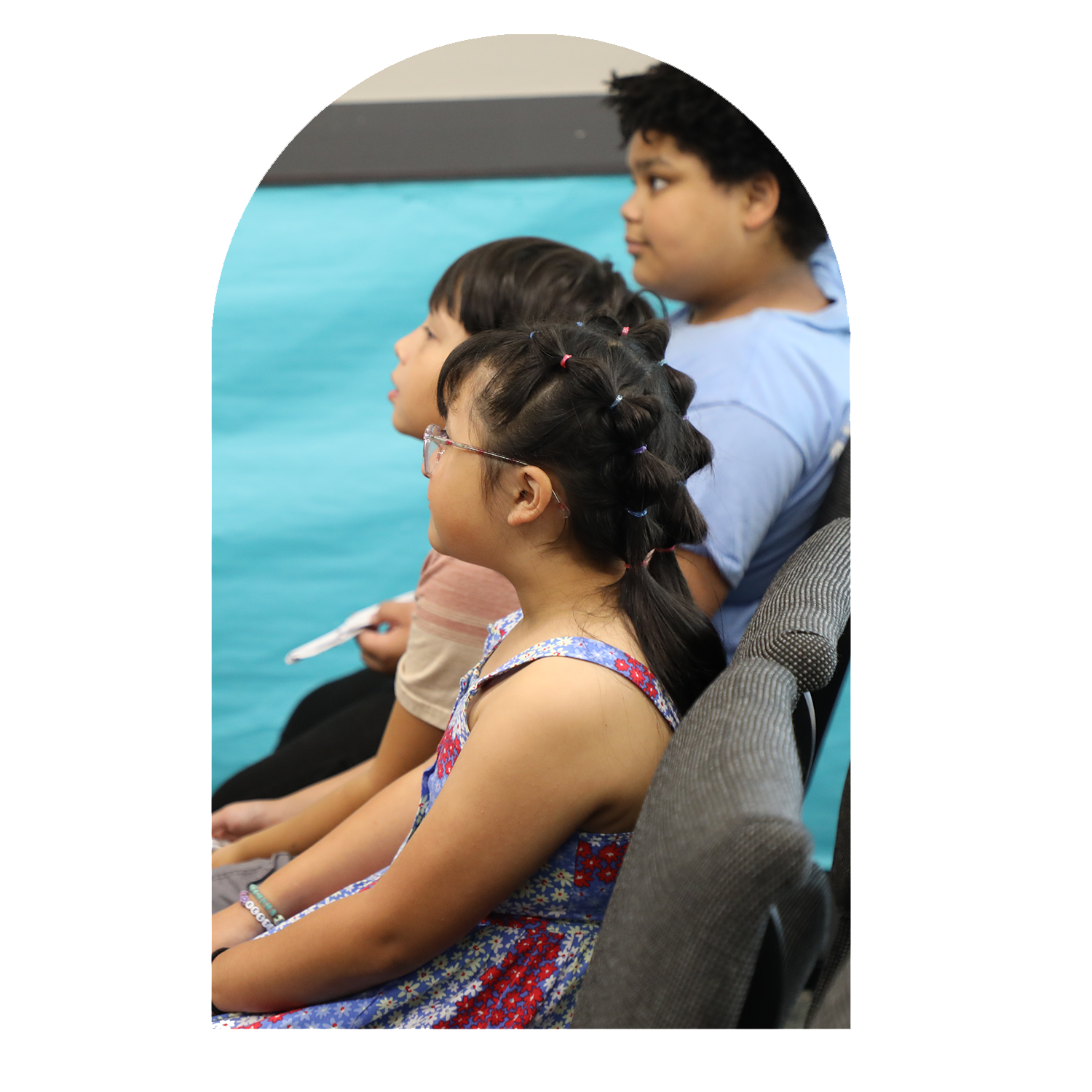 Group of four children sitting in a row, facing to the left, inside a room with a blue wall and gray chairs. The girl in the foreground has braided hair with colorful hair ties and wears glasses, a floral dress, and bracelets. Other children are boys with short hair, wearing casual clothing.
