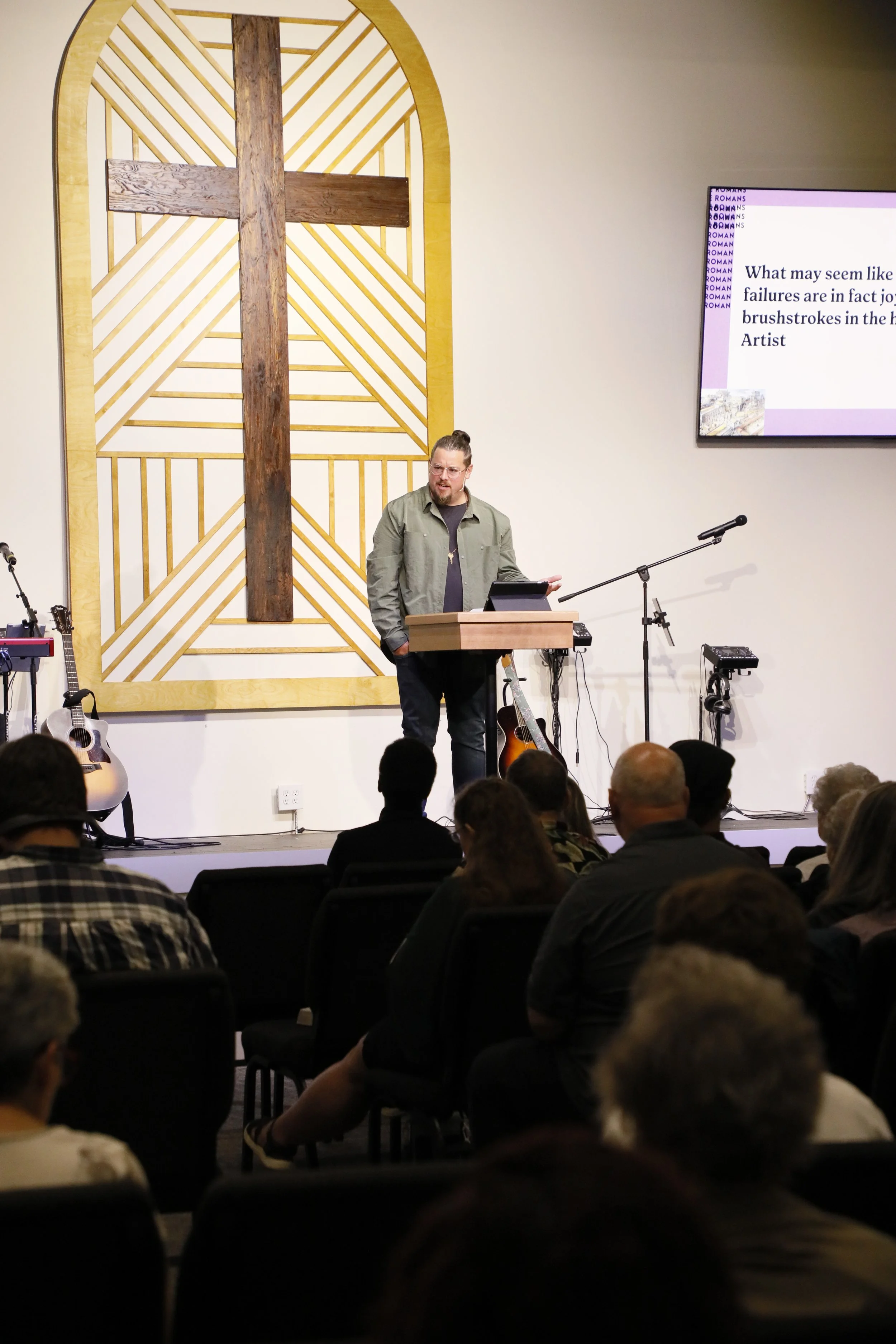 A man standing at a podium giving a presentation in a church or worship space, with a large decorative wooden cross on the wall behind him, and an electronic guitar on a stand to his left.