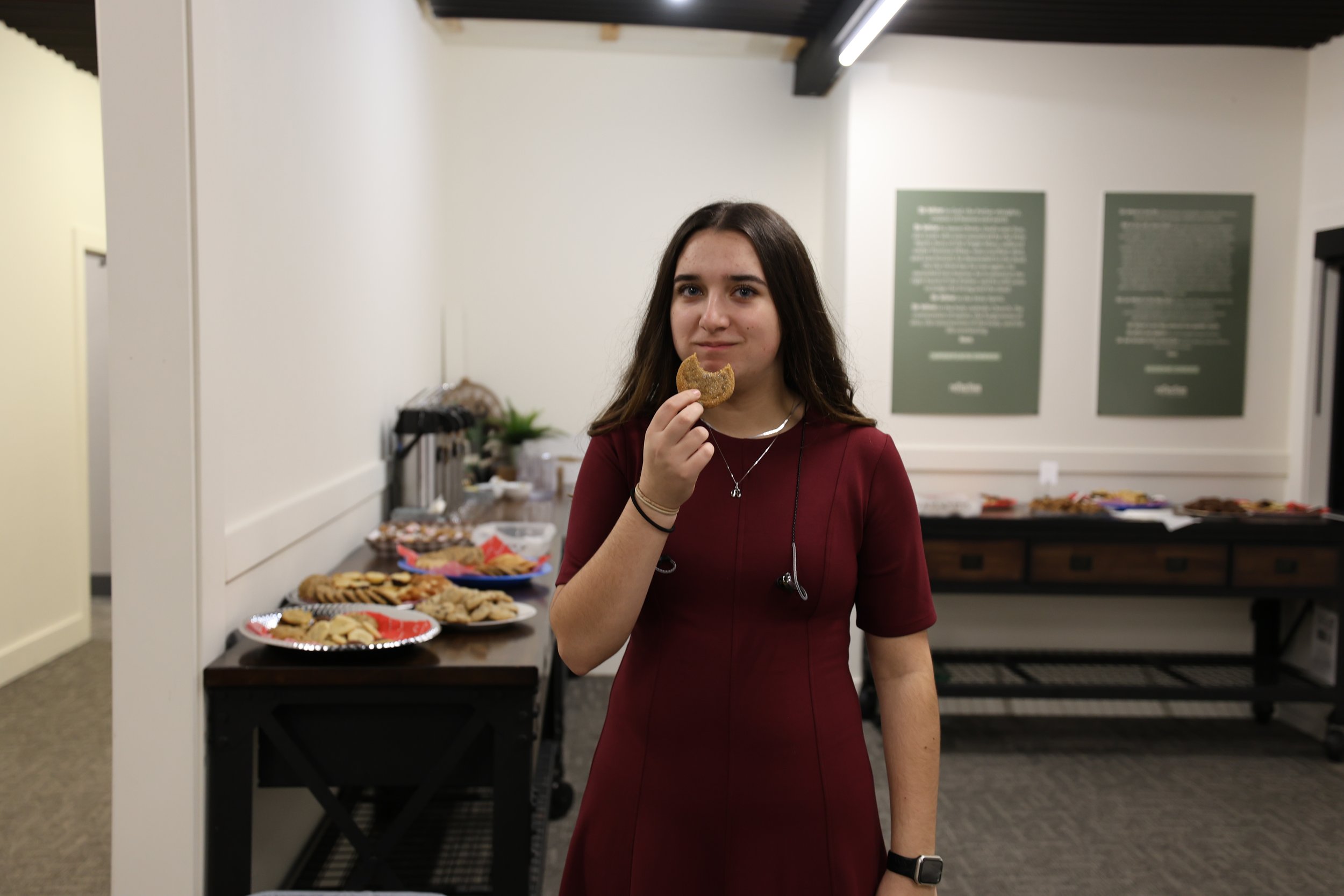 A young woman with long brown hair wearing a red dress and black watch is holding a cookie near her mouth. She is standing in front of a table with various cookies and snacks in a room with white walls, two green framed posters on the wall, and a serving table in the background.