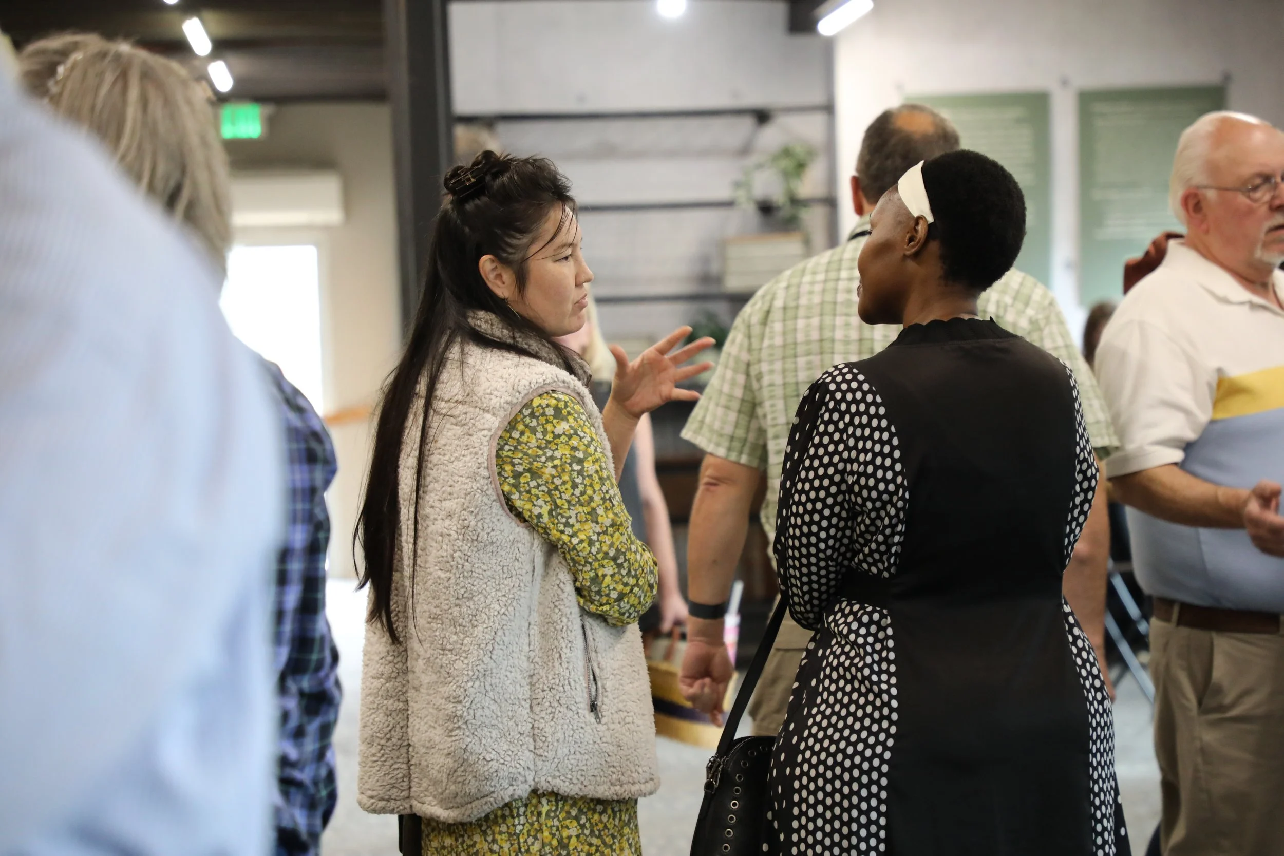 Two women in conversation, one with long dark hair wearing a cream fleece vest and floral dress, the other with short black hair wearing a black dress with white polka dots, in a crowded indoor setting.