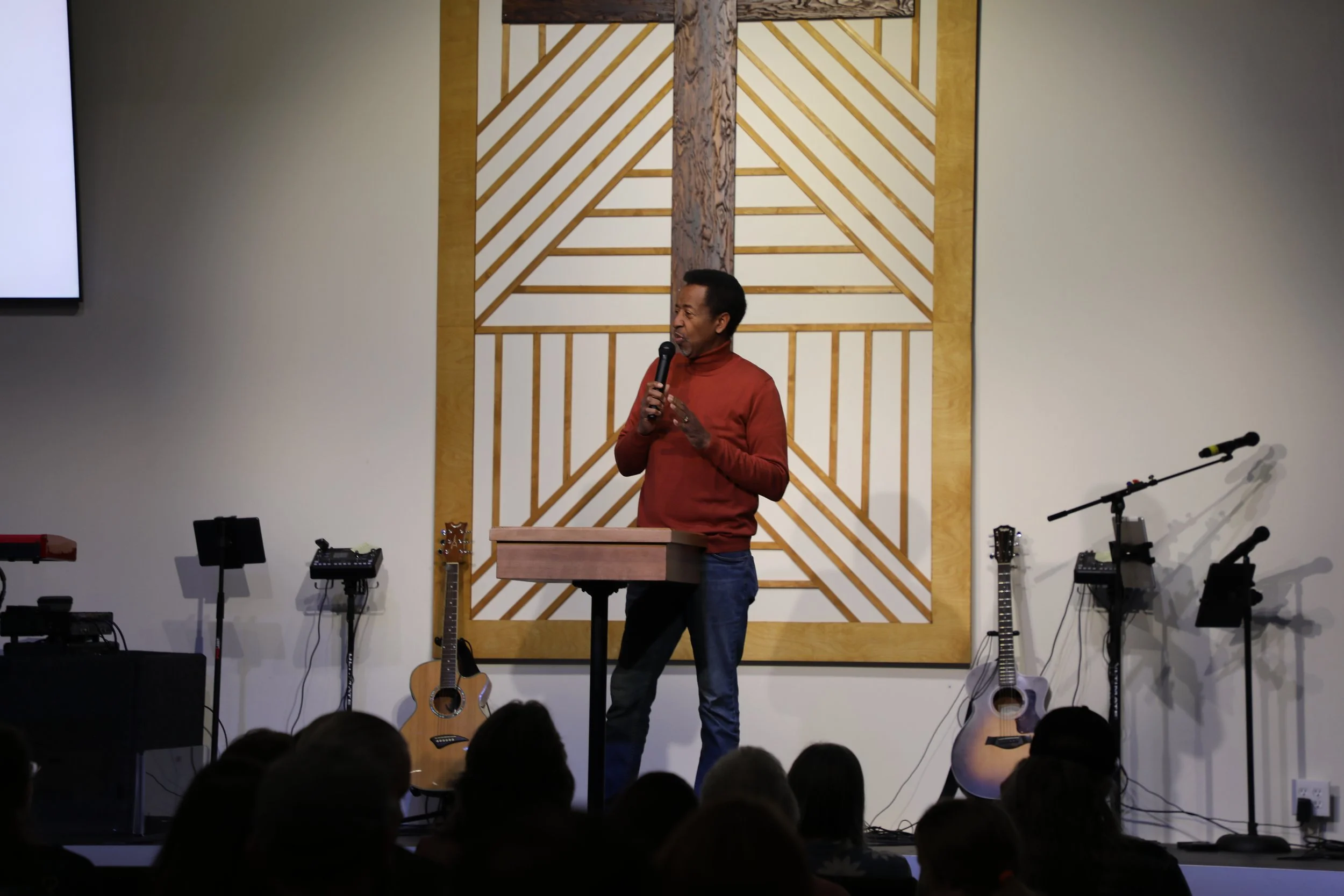 Man speaking into a microphone at a podium on stage, with musical instruments including guitars and microphones behind him, in a room with an audience.