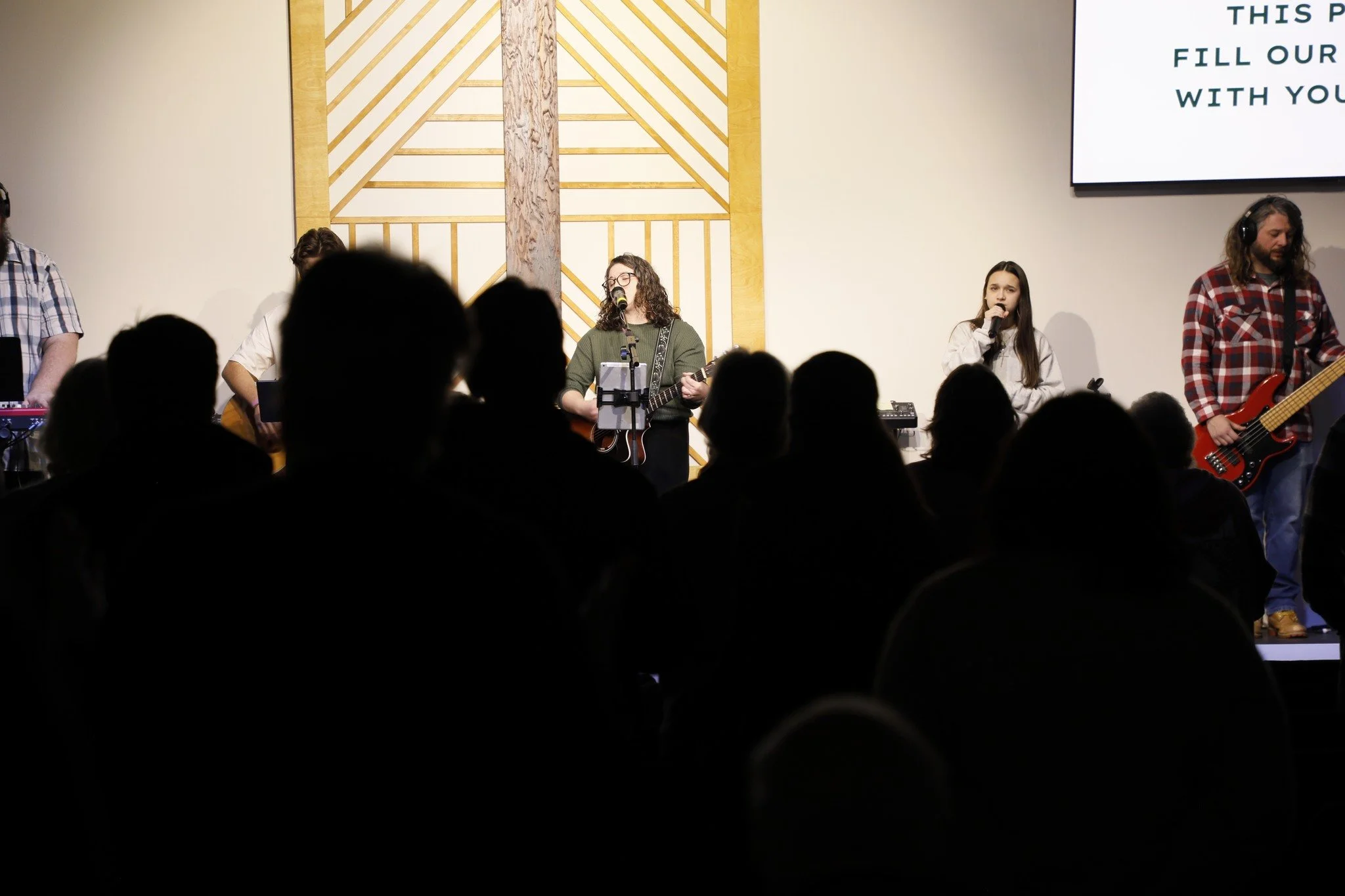 A band performing on stage with audience silhouettes in the foreground, in a room with a decorative wooden wall.