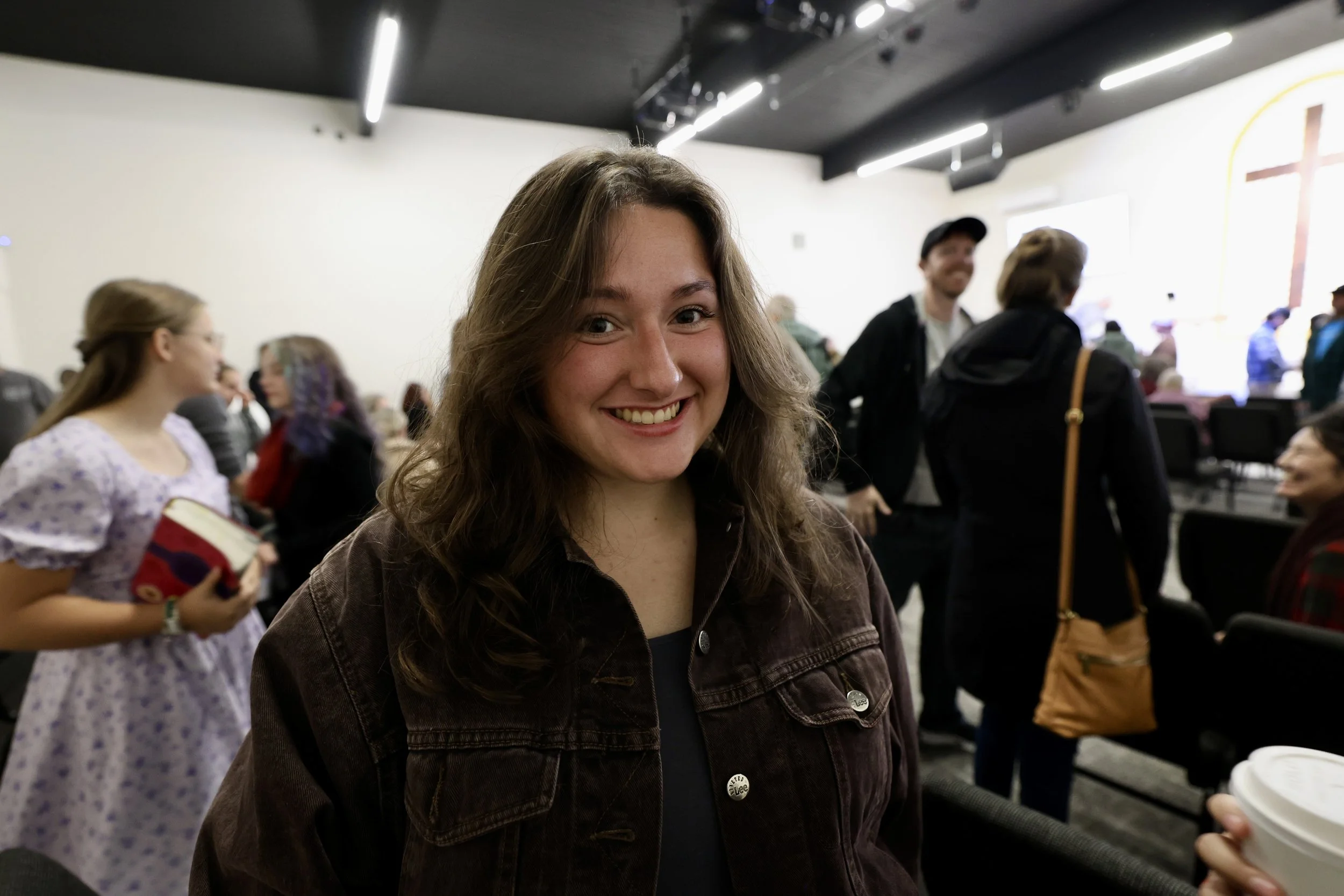 A smiling young woman with wavy brown hair standing in a crowded indoor space. She is wearing a brown denim jacket and is holding a white coffee cup. The background shows other people, some engaged in conversation, and a woman holding a book.