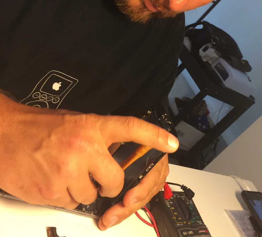 A person with a beard, wearing a black shirt with a graphic of an old cell phone and an Apple logo, is repairing a smartphone using a screwdriver. There is a digital multimeter connected with red and black probes on the table nearby, and a smartphone is visible in the background.