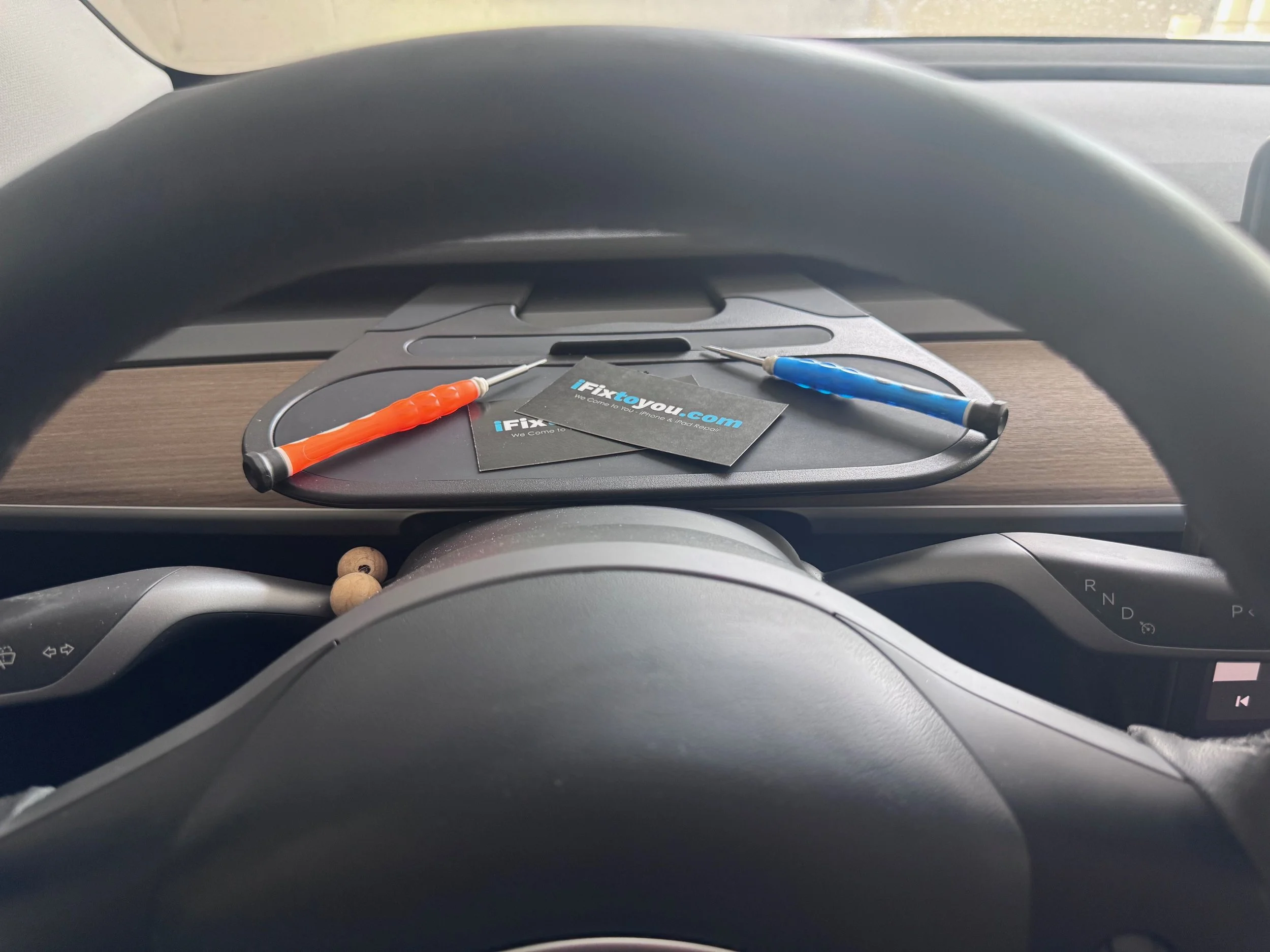 Inside a car, view from the driver's seat showing a steering wheel, dashboard, and a selling tray on top of the dashboard. The selling tray holds a blue and an orange screwdriver, and business cards for 'iFixToYou.com'.
